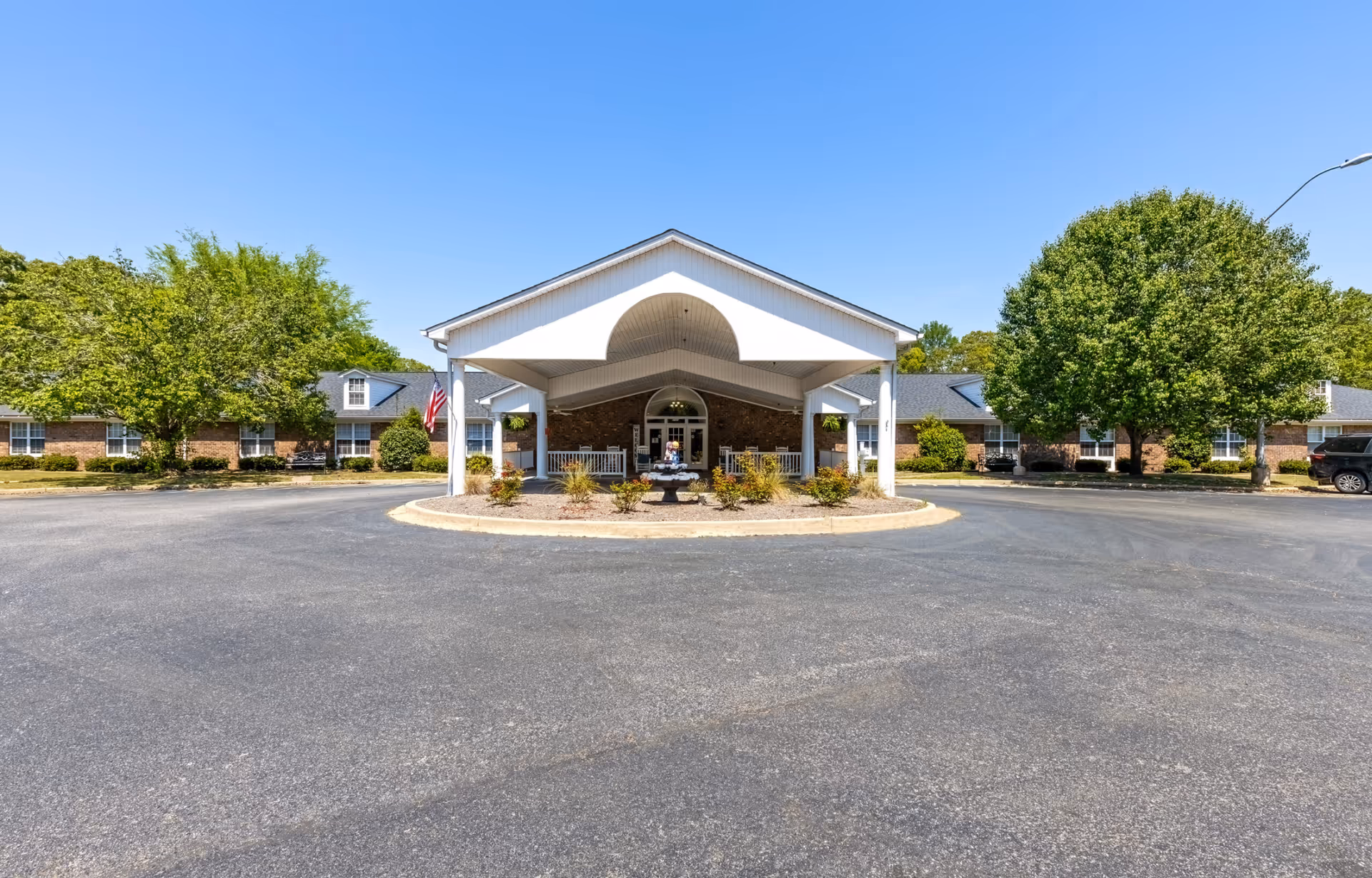 Front exterior view of The Gardens at Sumter facility featuring a covered entrance with white pillars, a circular driveway, landscaped bushes, and large green trees under a clear blue sky.