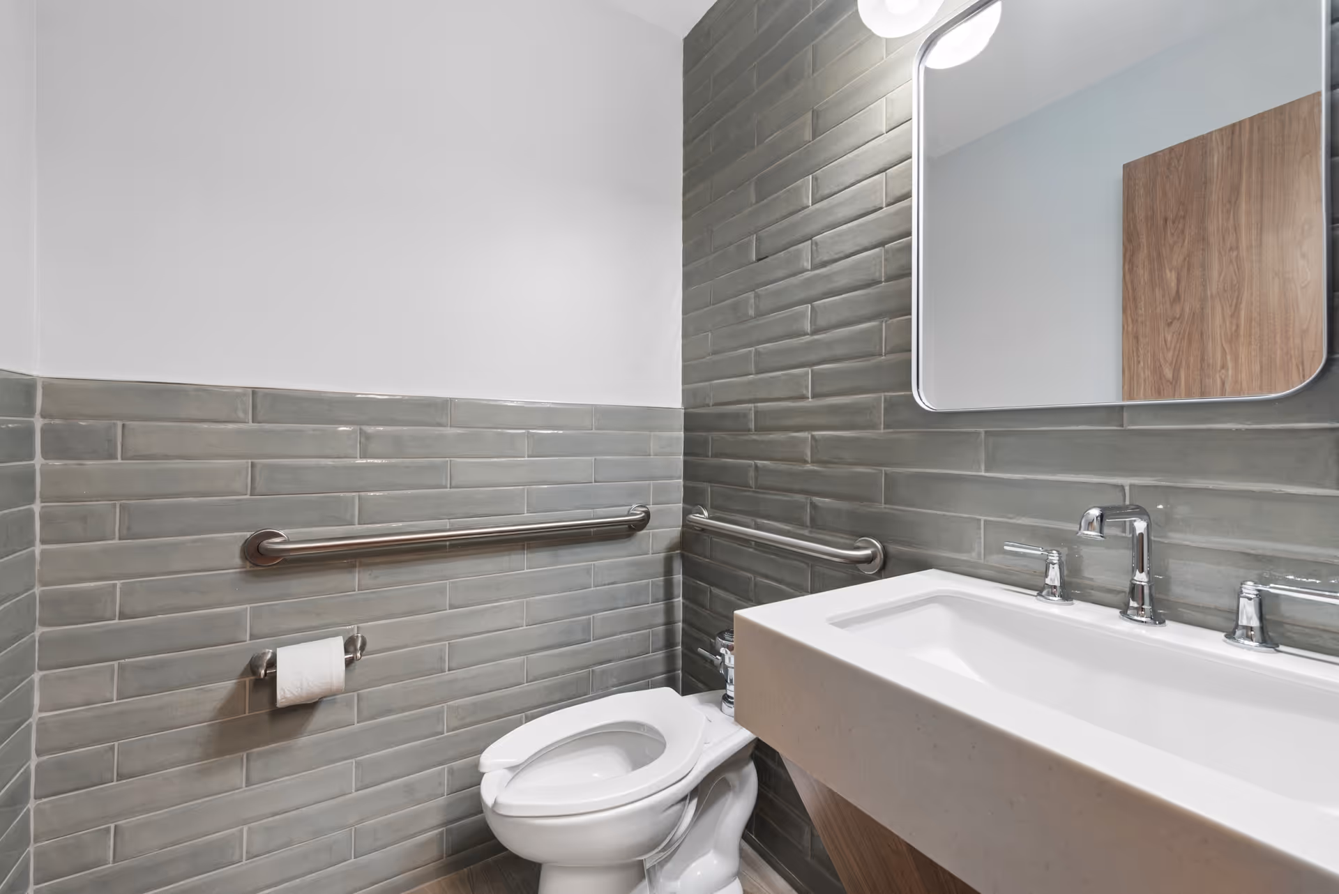 Accessible bathroom with gray tile, grab bars, toilet, and a sink with mirror.