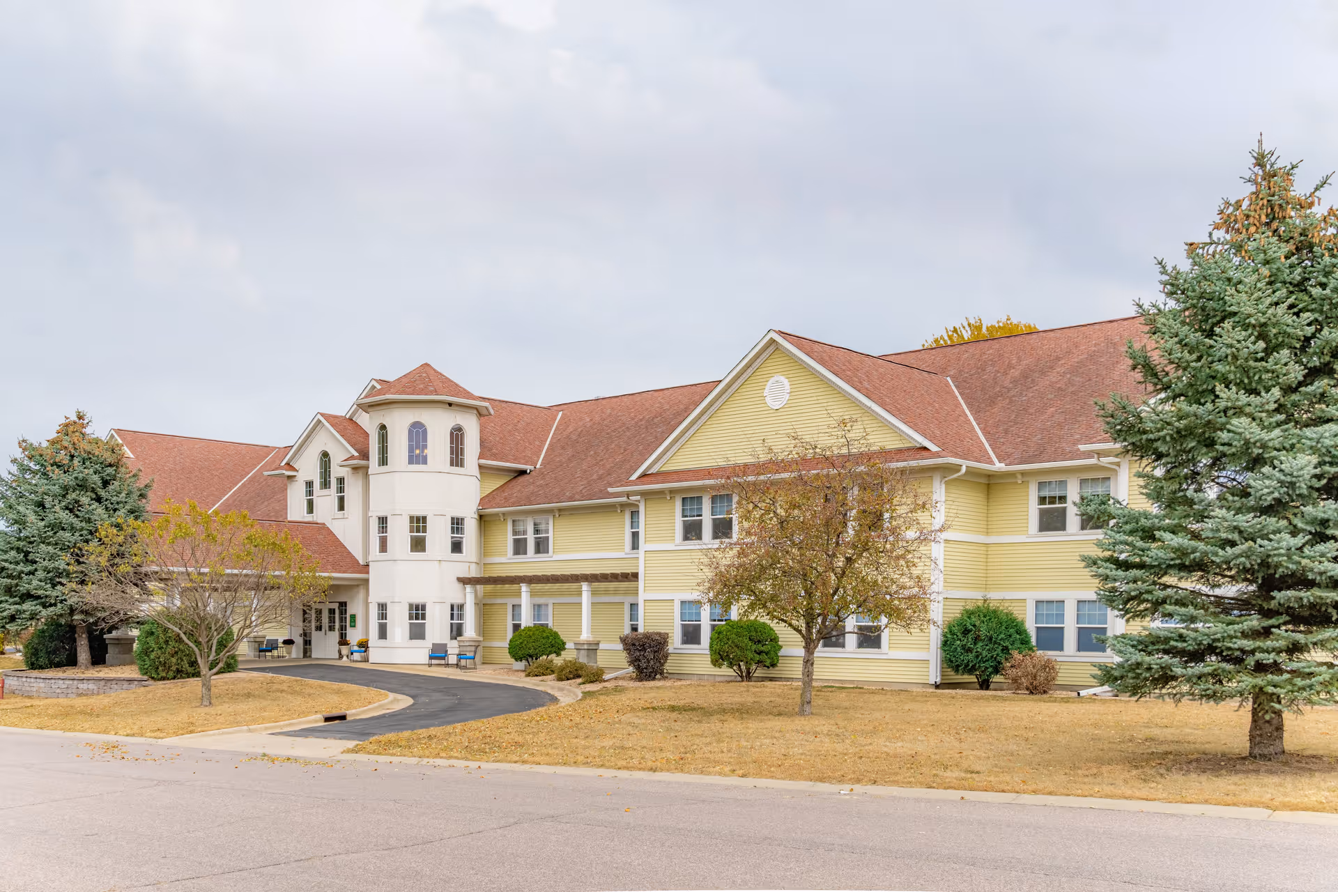 Exterior view of a two-story senior living facility with yellow siding and a red roof, surrounded by trees and a paved driveway under a cloudy sky.
