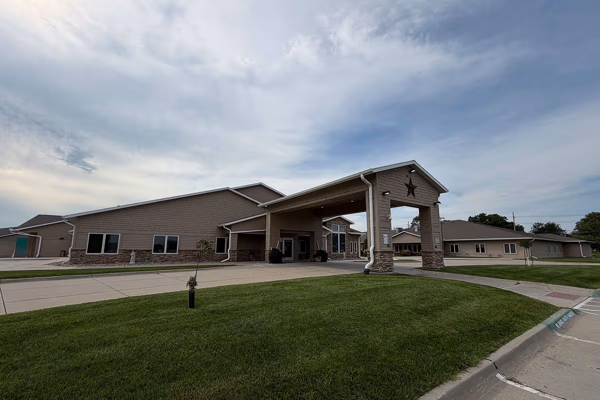 Single-story senior living facility with covered entrance and star emblem, surrounded by green lawn.
