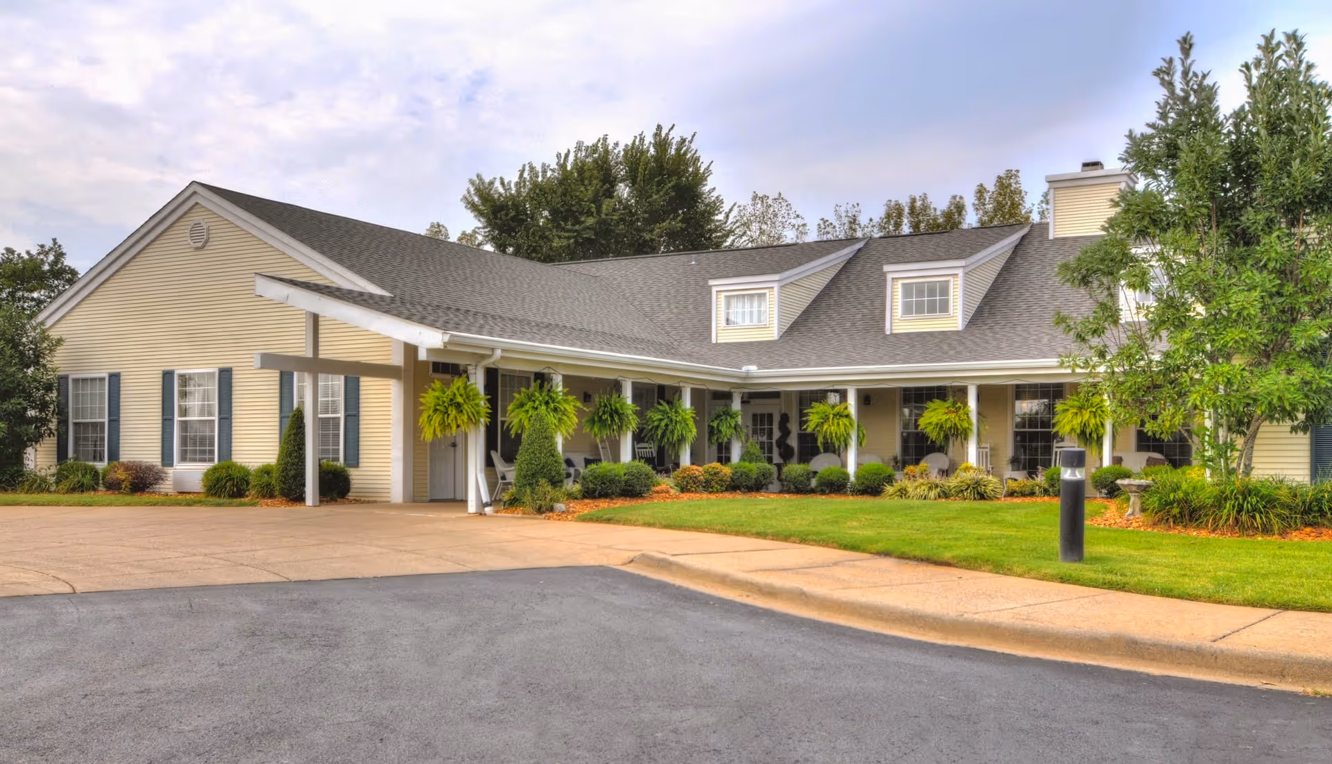 Front entrance of a single-story residential building with a covered porch, hanging fern planters, and a manicured lawn and driveway.