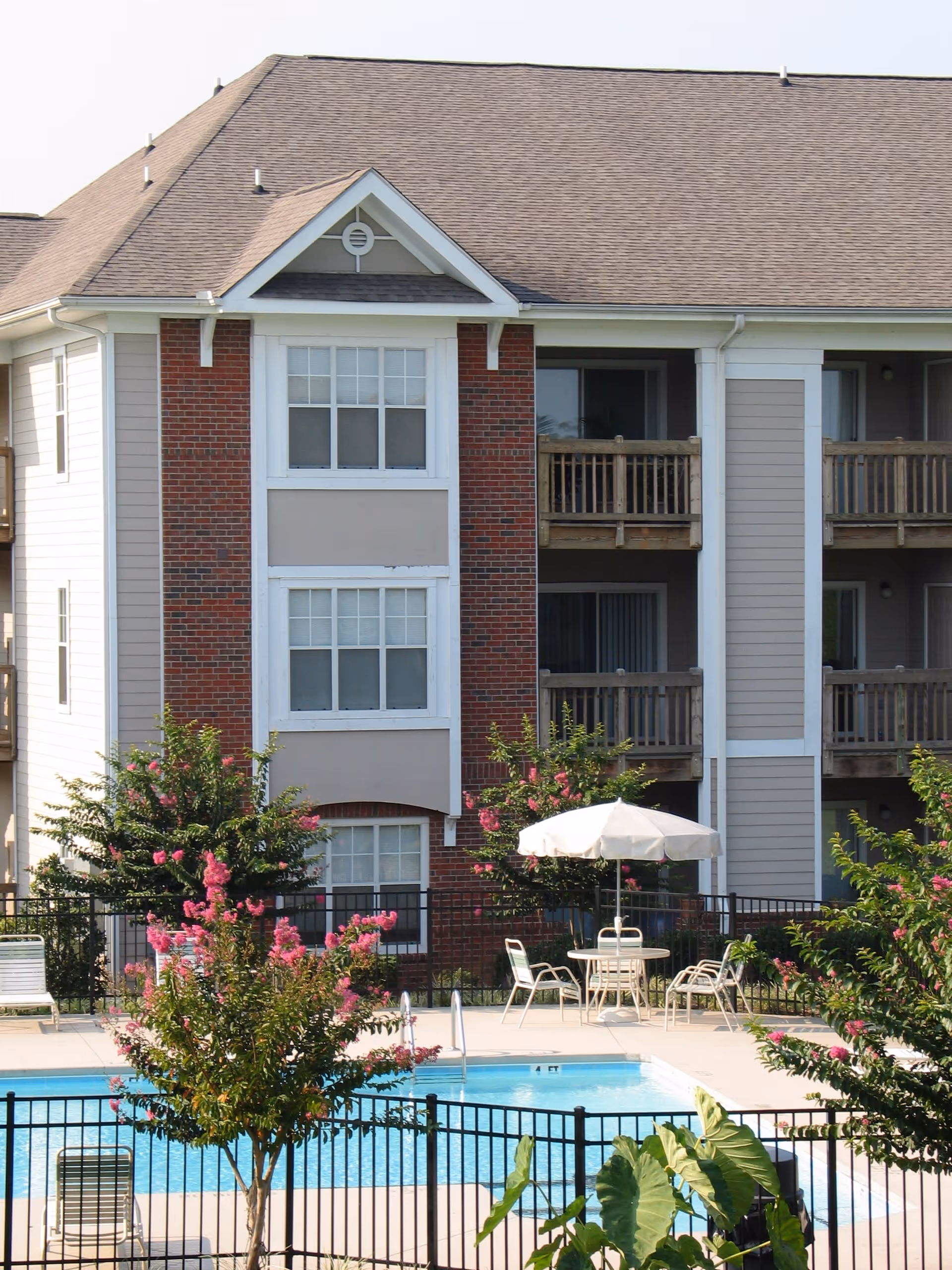 Outdoor view of Riverwalk Apartments showing a swimming pool surrounded by a black metal fence, patio chairs, a table with a white umbrella, flowering trees, and a three-story building with balconies in the background.