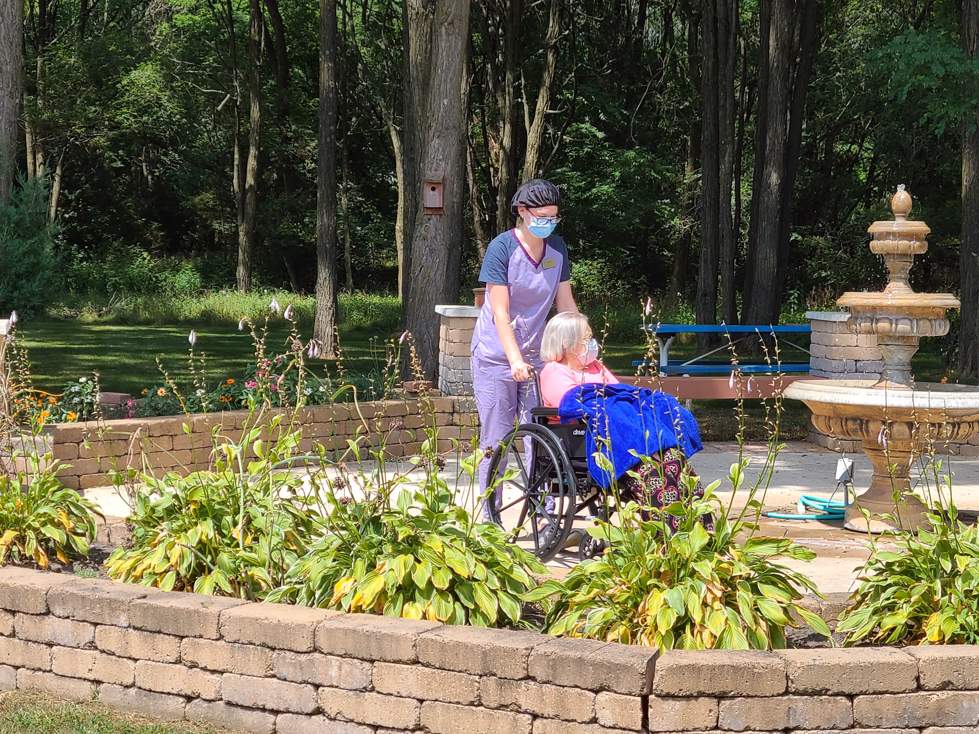 A caregiver wearing a mask and purple scrubs is pushing an elderly woman in a wheelchair outdoors near a stone fountain. The elderly woman is covered with a blue blanket. They are surrounded by greenery and plants with trees in the background.