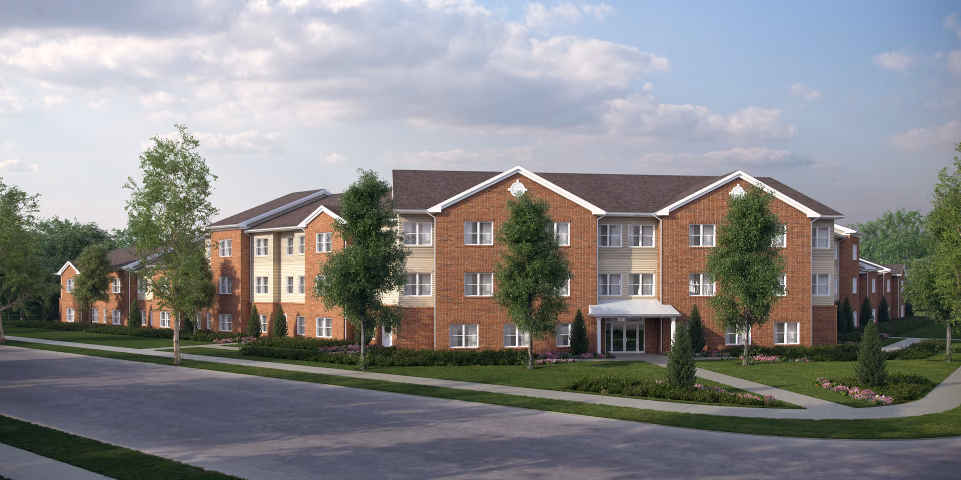 Exterior view of a three-story senior living residence building with a combination of red brick and beige siding. The building is surrounded by green lawns, trees, and landscaped flower beds under a partly cloudy sky.