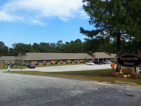Exterior view of Fayetteville Manor, a single-story brick building with a parking lot in front. Several cars are parked, and there are trees surrounding the area under a partly cloudy sky.