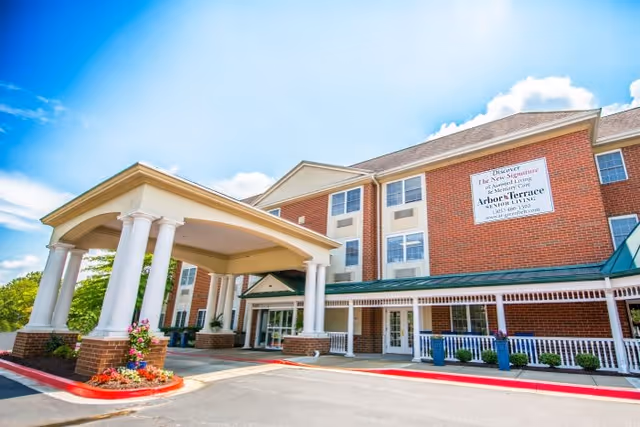Exterior view of Arbor Terrace Senior Living facility showing a large brick building with white columns at the entrance, a covered driveway, and a sign on the building advertising the senior living community.