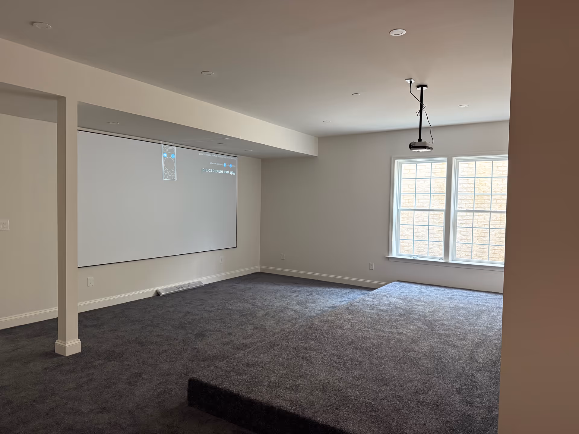 Empty recreation room with a projector screen, large window, and gray carpet.