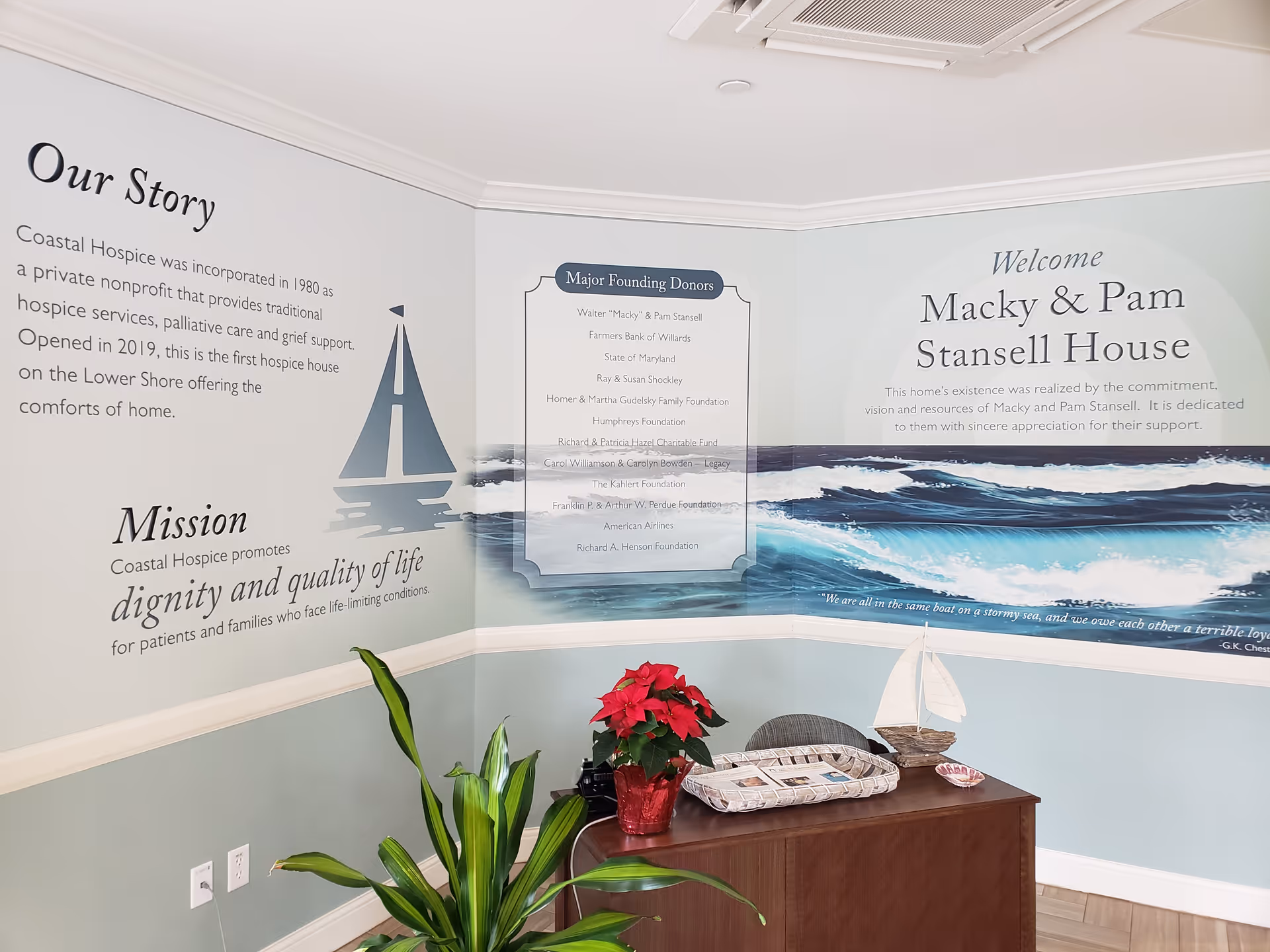 Interior corner with wall murals, welcome messages, desk, poinsettia, and decorative boats on a side table.