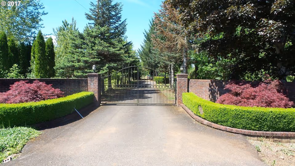 A gated entrance to a property with a black metal gate flanked by brick pillars and walls. The driveway leads through the gate, lined with neatly trimmed green hedges and red-leafed bushes. Tall trees and greenery surround the area under a clear blue sky.