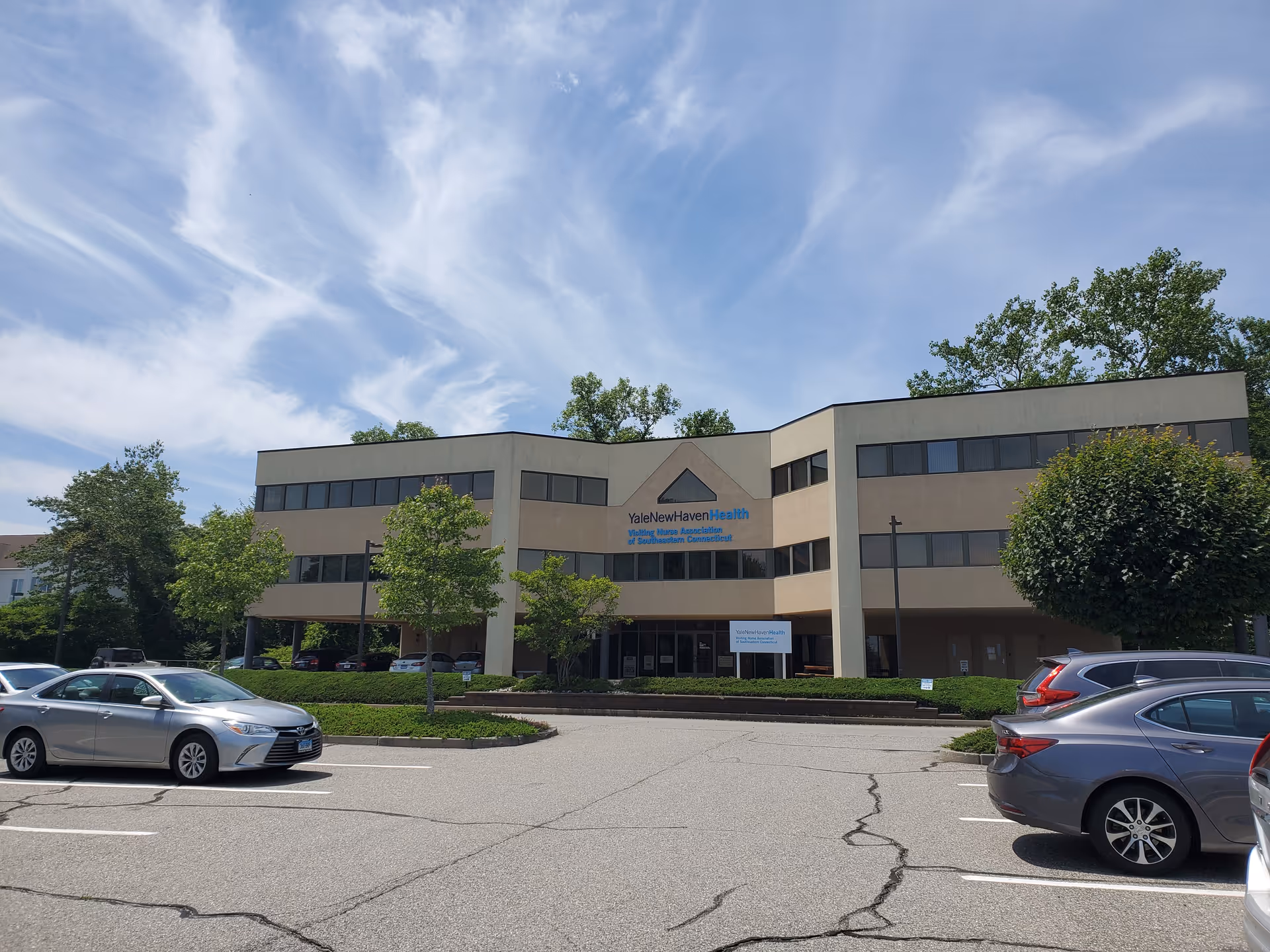 A beige medical building with a front entrance, parking lot, and green trees.