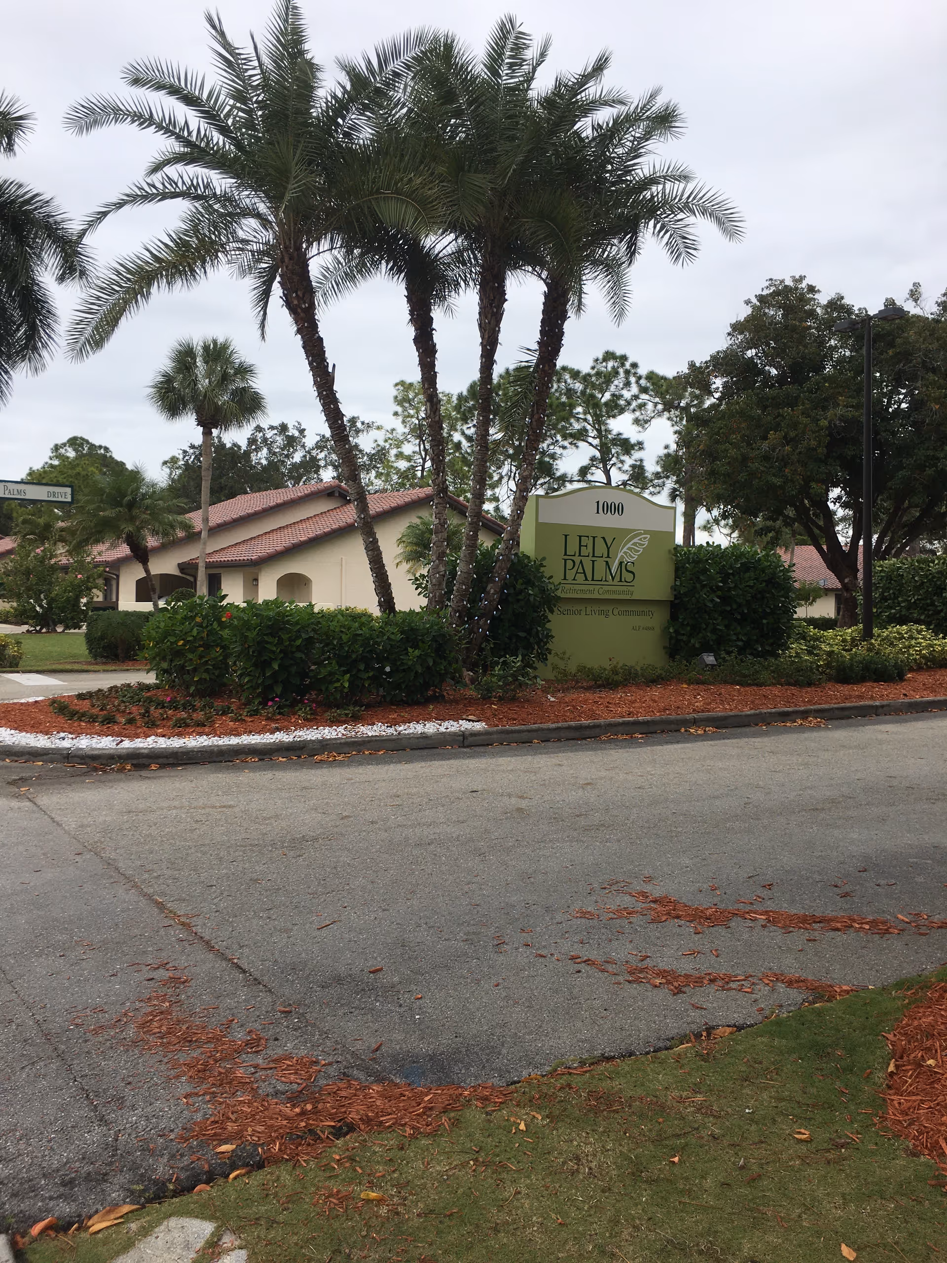 Exterior view of Lely Palms Senior Living Community with palm trees and a sign
