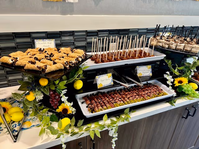 A buffet table with trays of assorted desserts including chocolate chip cookies, cake pops, and other bite-sized treats. The table is decorated with artificial greenery, lemons, grapes, and sunflowers, creating a fresh and inviting presentation.