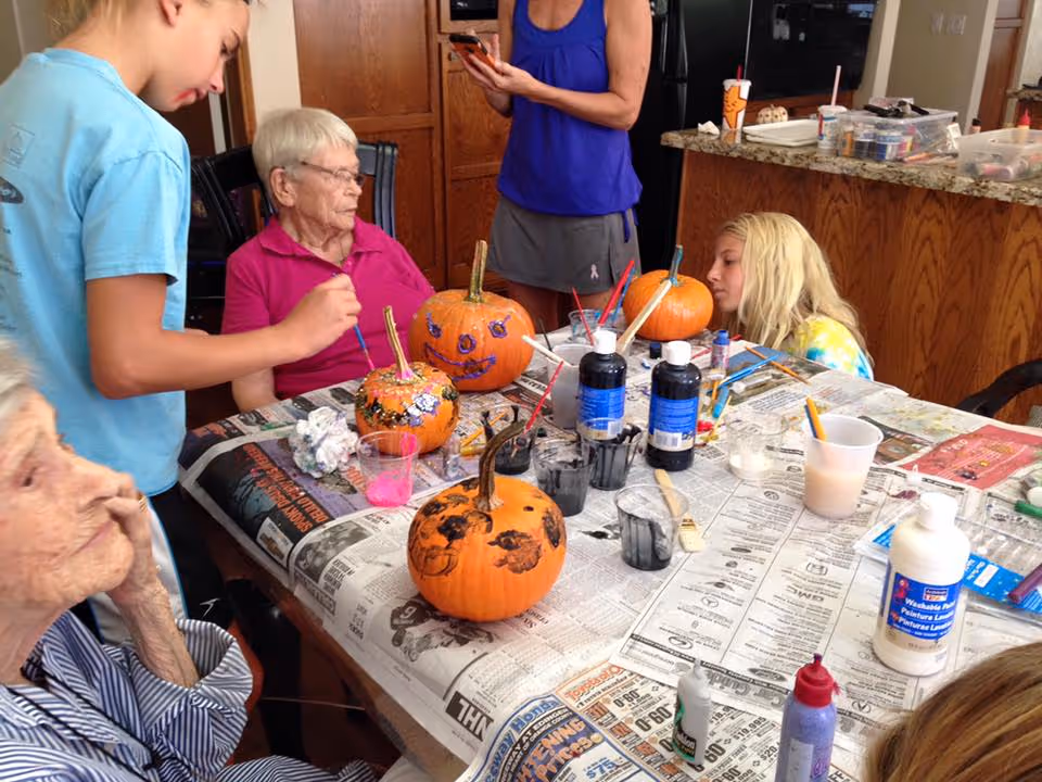 A group of elderly women and younger individuals sitting and standing around a table covered with newspapers, painting and decorating small pumpkins with various colors of paint and brushes in a cozy indoor setting with wooden cabinets and a granite countertop in the background.