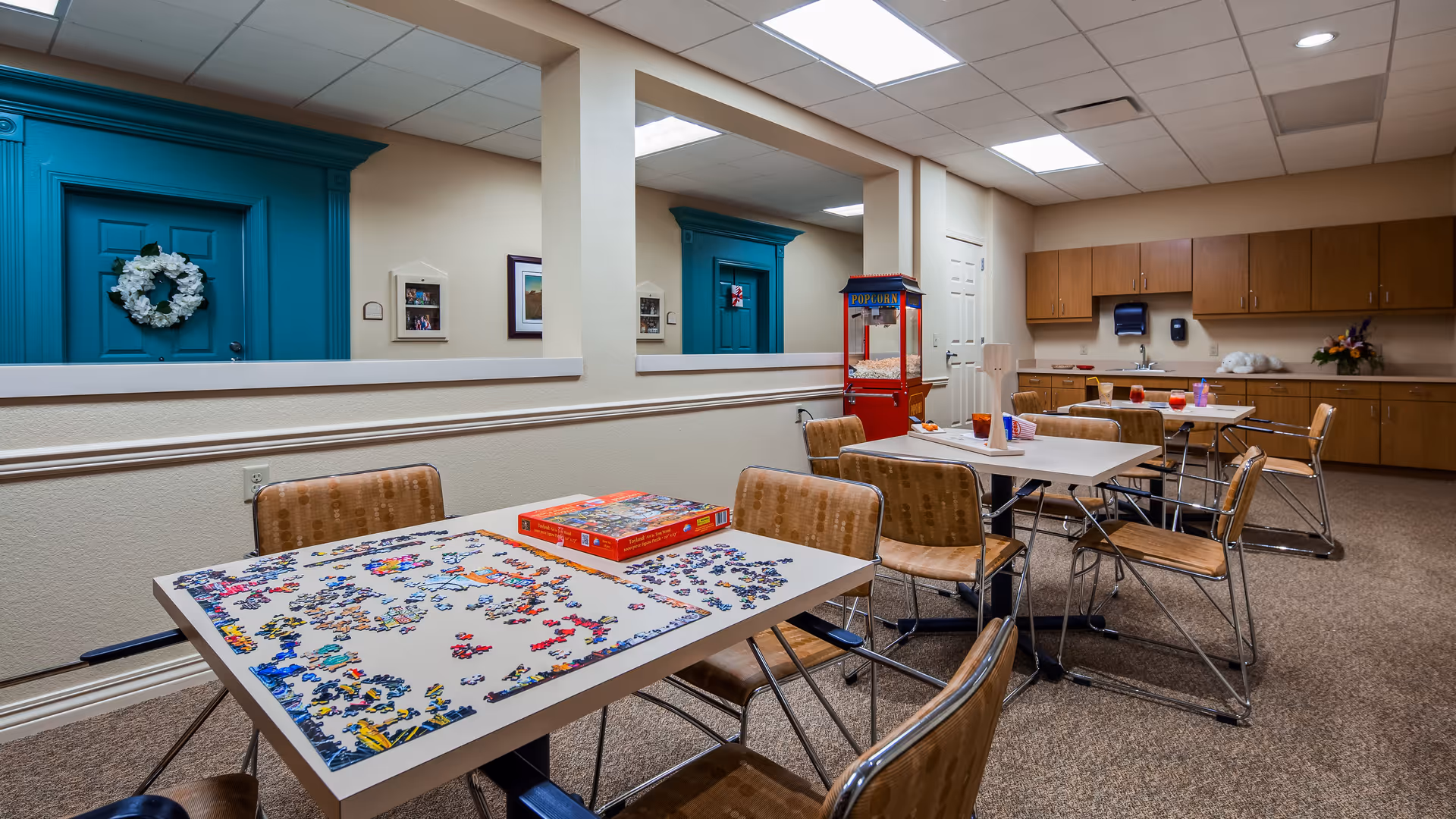 A well-lit activity room with tables and chairs arranged for group activities. One table has a partially completed jigsaw puzzle with the puzzle box on top. In the background, there is a popcorn machine and a kitchenette area with wooden cabinets, a sink, and paper towel dispensers. The walls are beige with teal door frames and decorative wreaths on the doors.