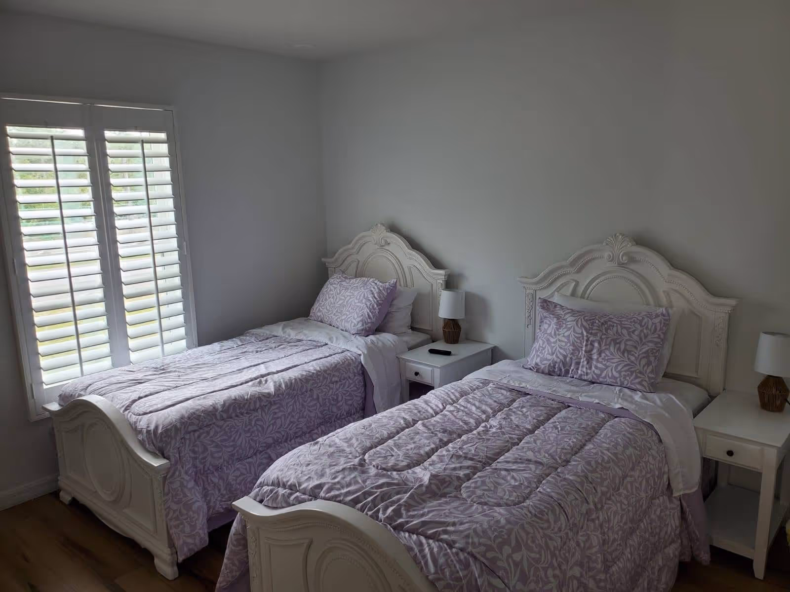 Two twin beds with ornate white headboards, lavender bedding, and nightstands in a bright bedroom.