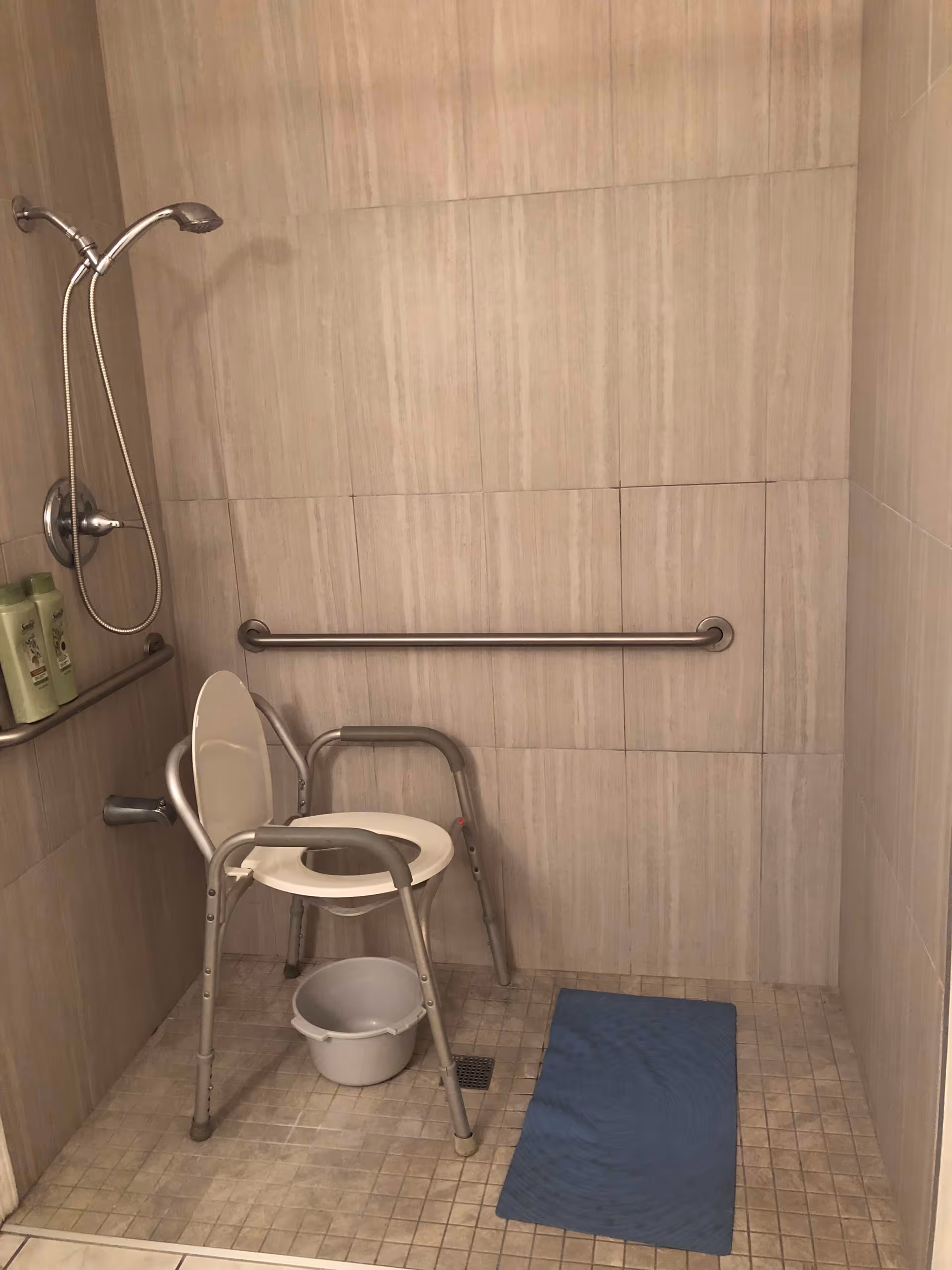 Accessible tiled shower with a handheld showerhead, wall grab bars, a commode shower chair and a blue bath mat.