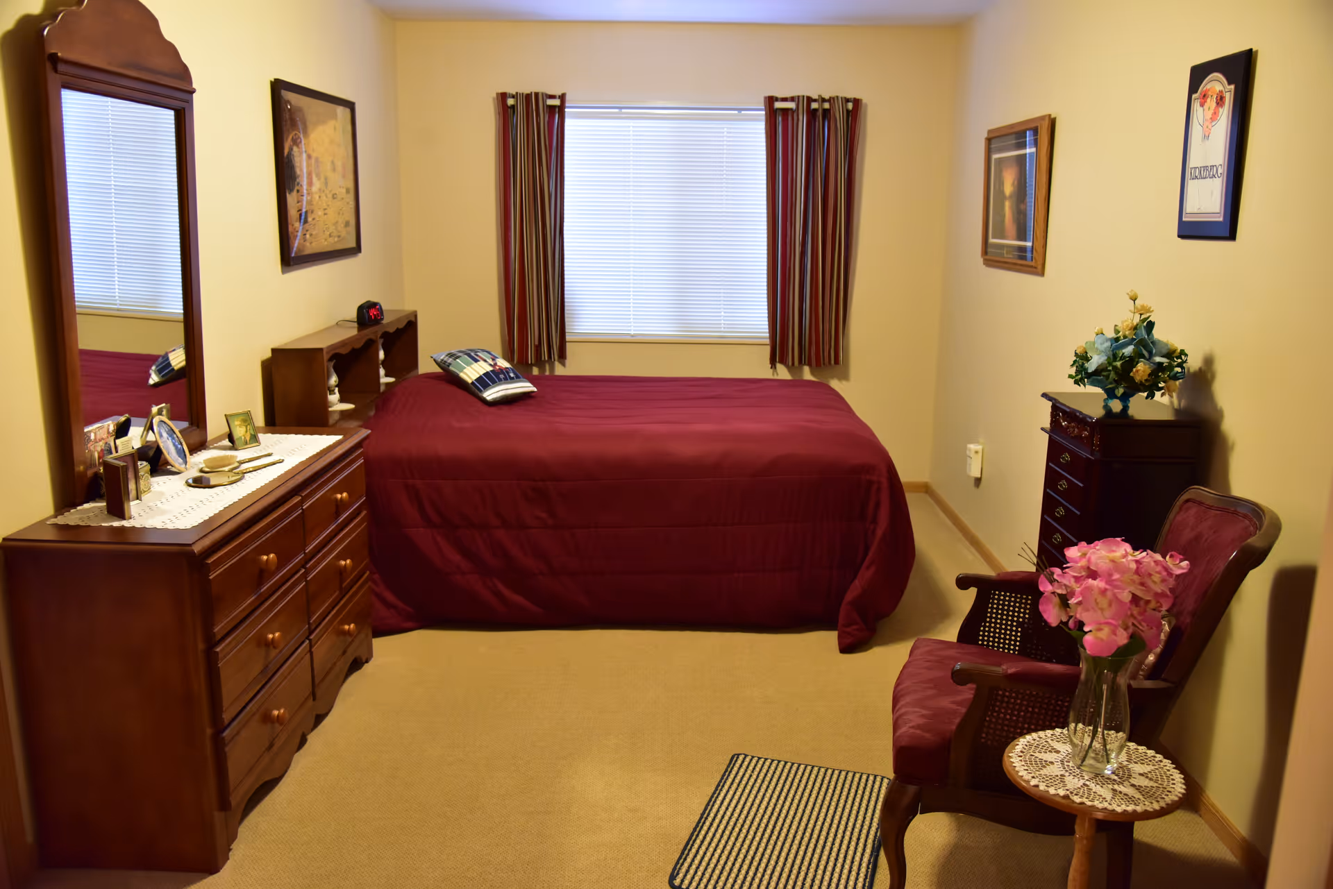 A bedroom with a bed covered in a maroon bedspread, a wooden dresser with a mirror on the left, a small wooden chest with a flower vase on the right, and a wooden chair with a pink cushion and a small round table with pink flowers in a vase. The window at the back has blinds and striped curtains.