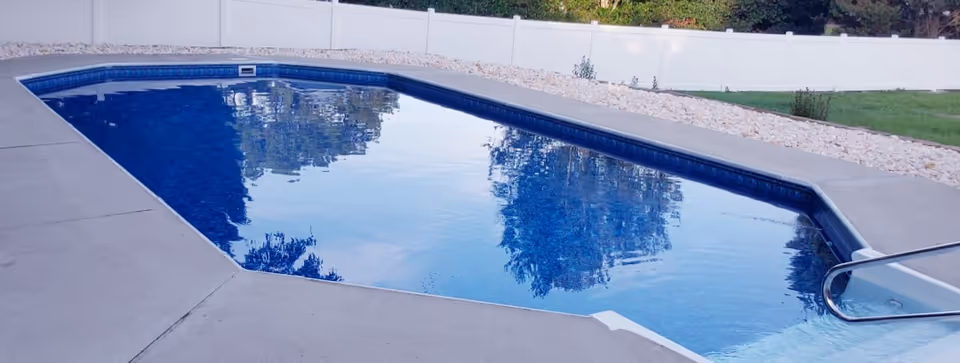 Blue rectangular swimming pool with concrete deck and white fence in a sunny backyard.