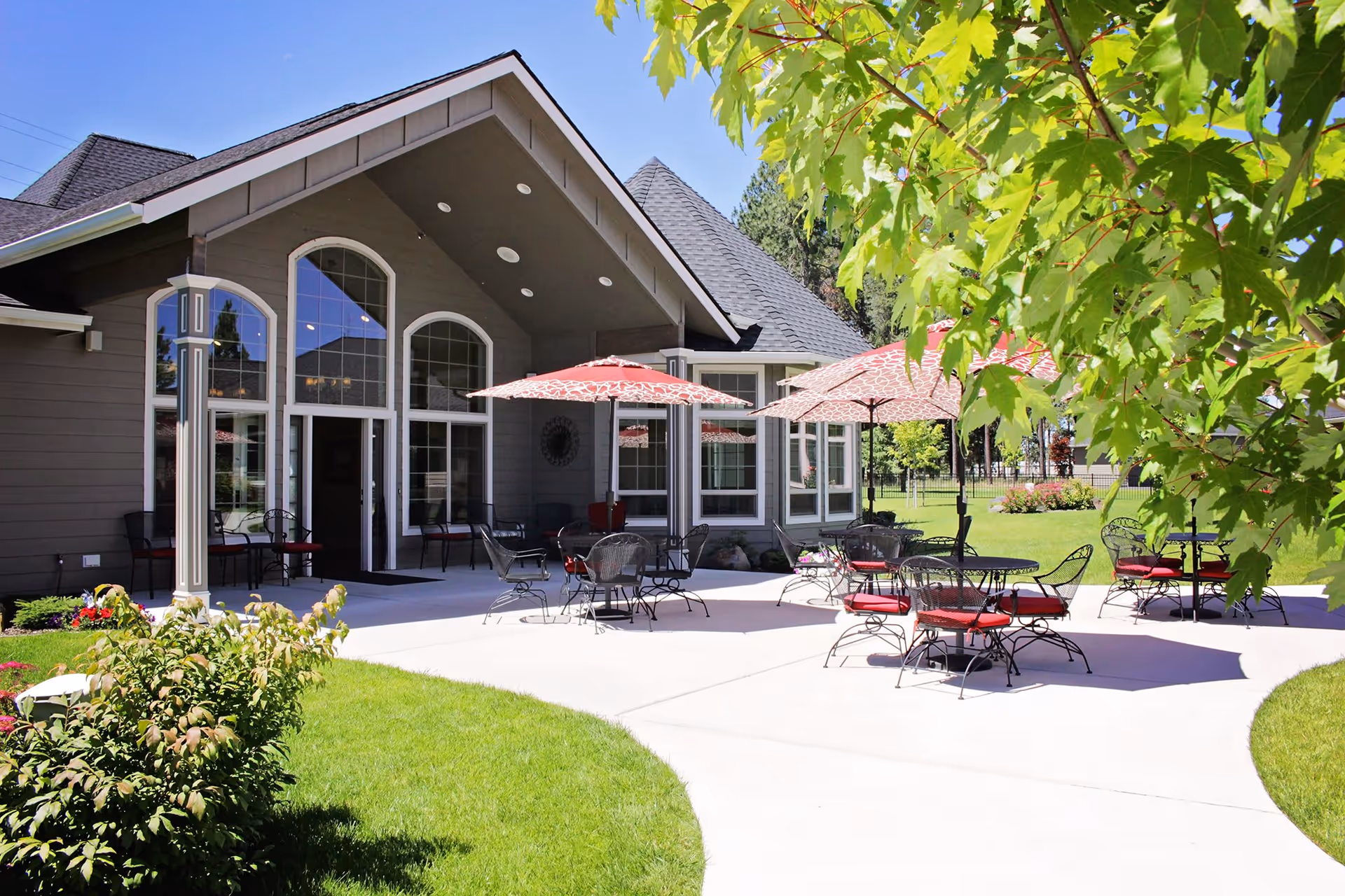 Outdoor patio with round tables, red umbrellas, and chairs in front of a senior living building.