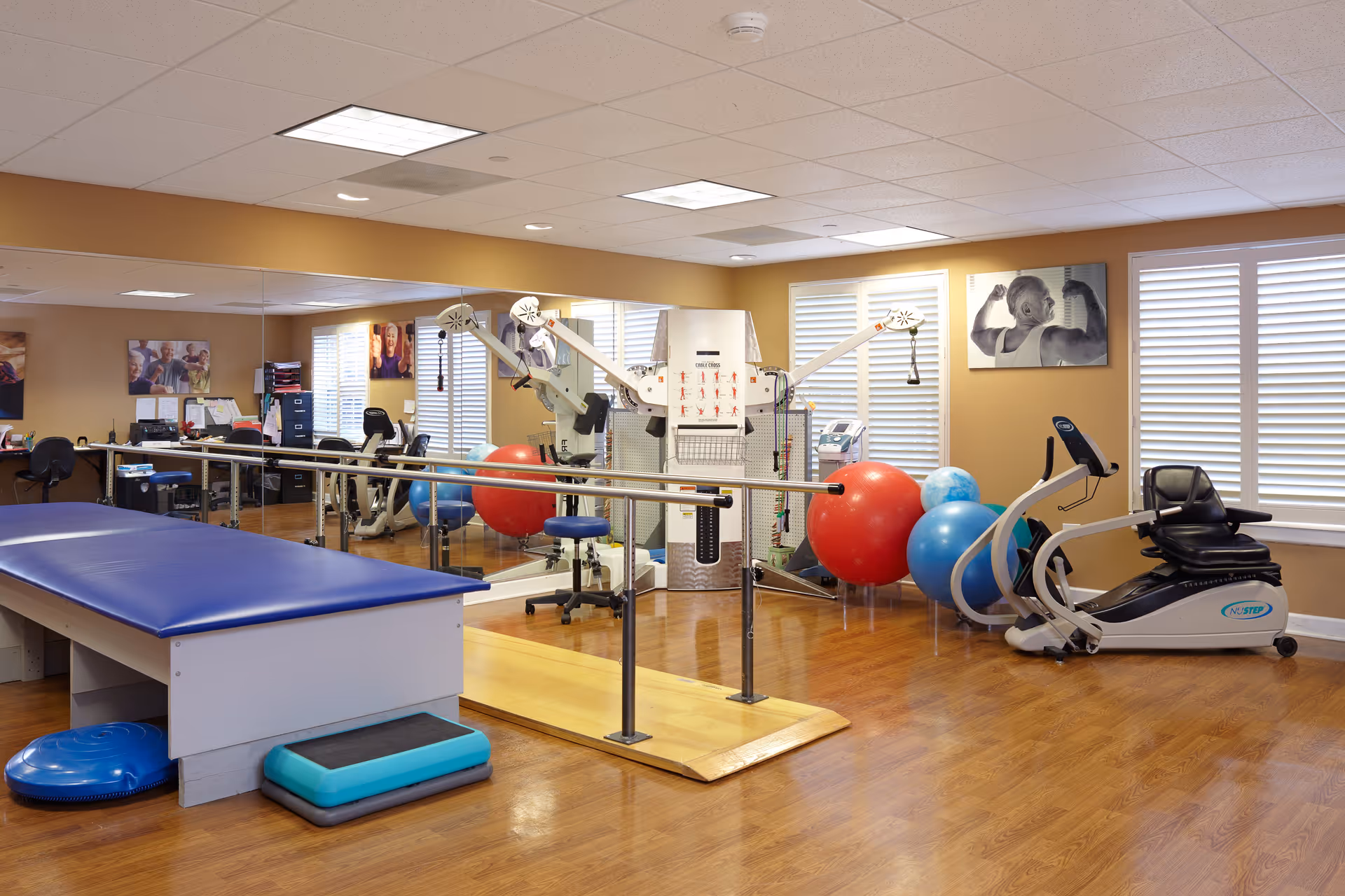 A senior living facility exercise room with wooden floors, a blue padded exercise table, parallel bars for walking support, various exercise balls, a recumbent exercise bike, and a multi-functional exercise machine. The room has large windows with white blinds and motivational fitness posters on the walls.