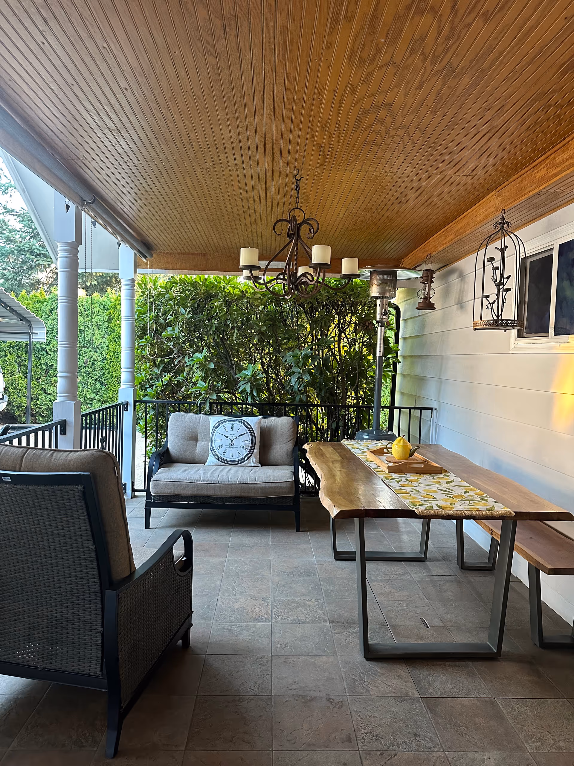 Covered outdoor patio with seating, a wooden table, chairs, and lush greenery.