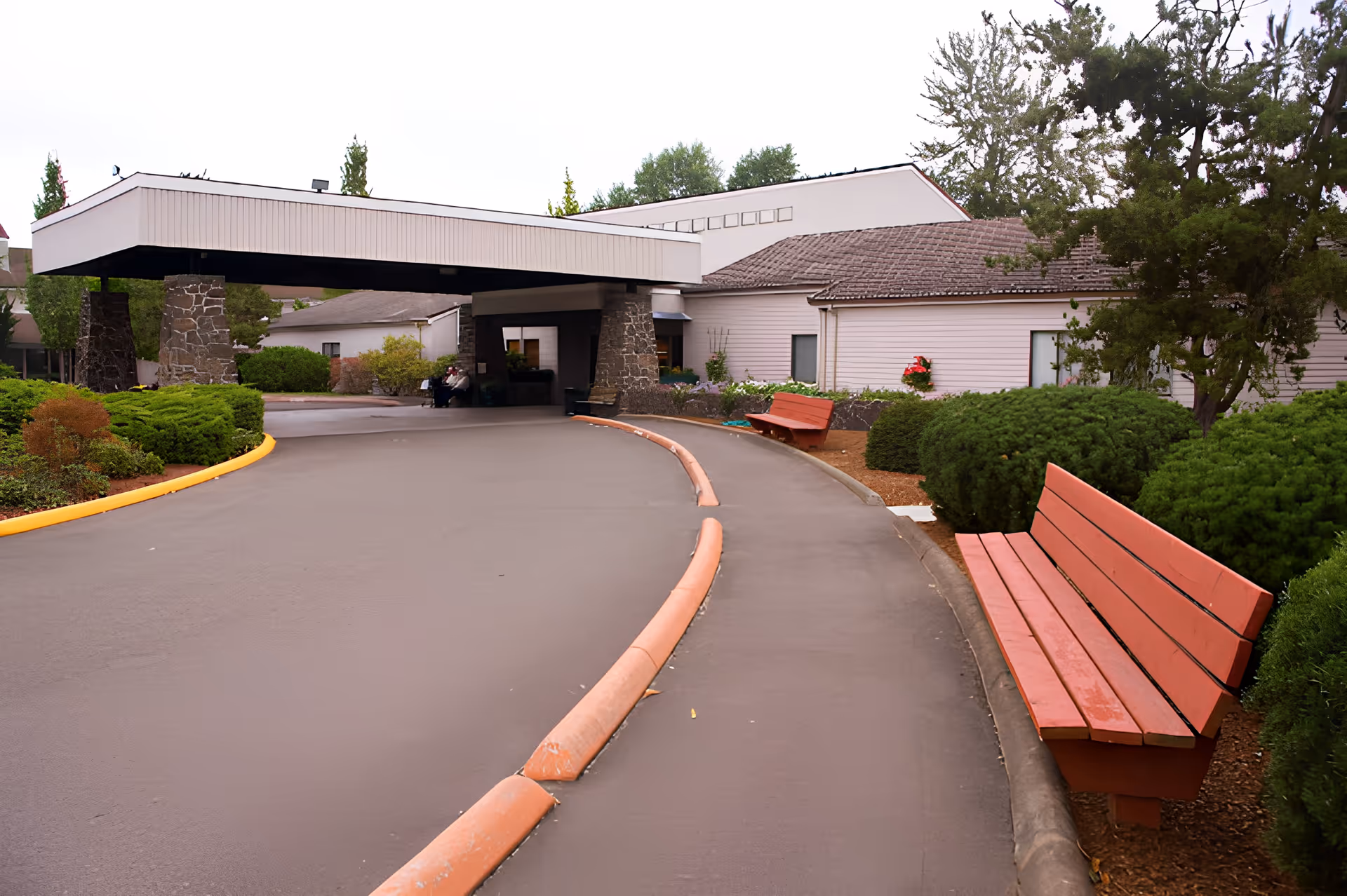 Entrance driveway of Churchill Estates Retirement Community with a covered drop-off area supported by stone pillars. There are red benches along the sidewalk and well-maintained bushes and trees surrounding the building.