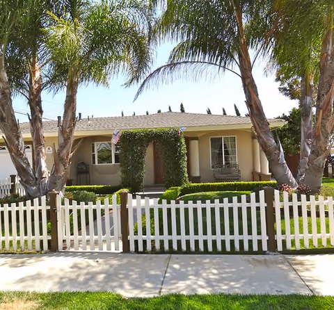 Front view of Windham House 1 with a white picket fence and palm trees.
