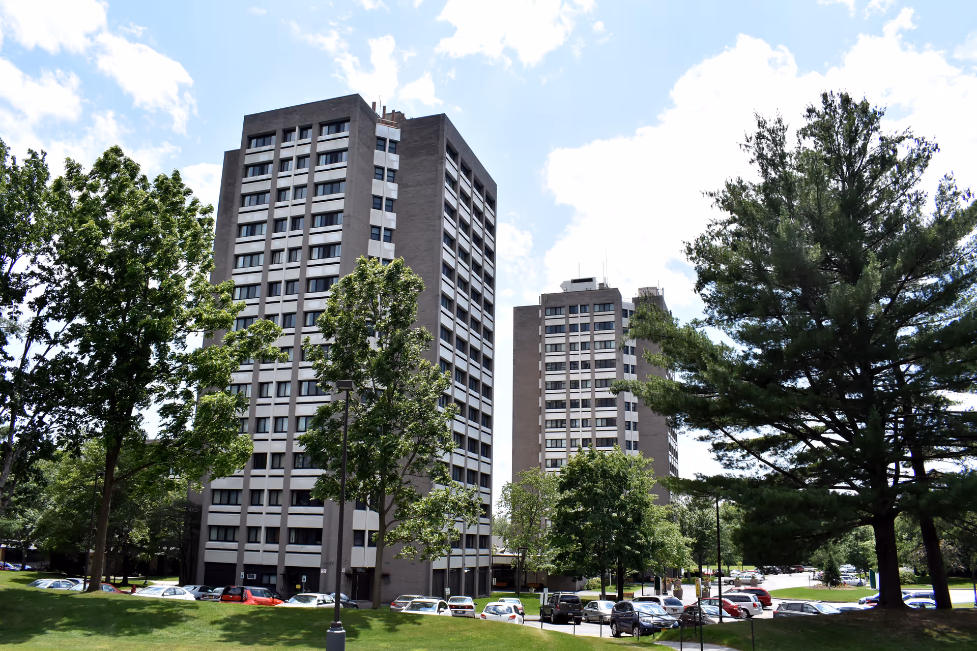 Two tall apartment buildings surrounded by green trees and a parking lot with several cars parked. The sky is partly cloudy with blue patches visible.