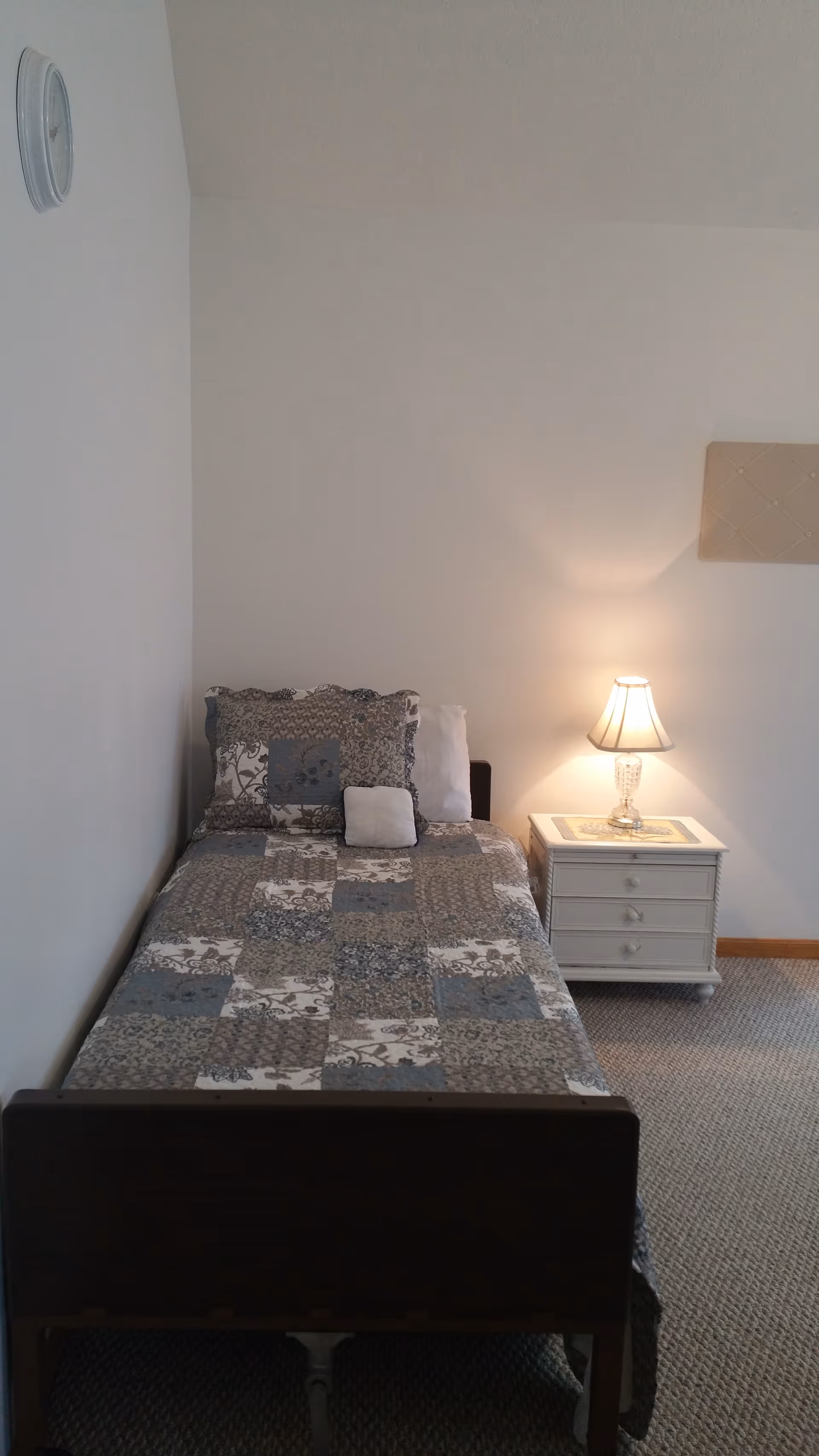 A small bedroom with a single bed, quilt, and nightstand with lamp.