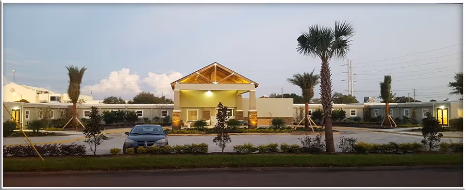 Exterior view of Walton Place Assisted Living facility at dusk, showing a single-story building with a covered entrance, palm trees, shrubs, and a parked car in front.