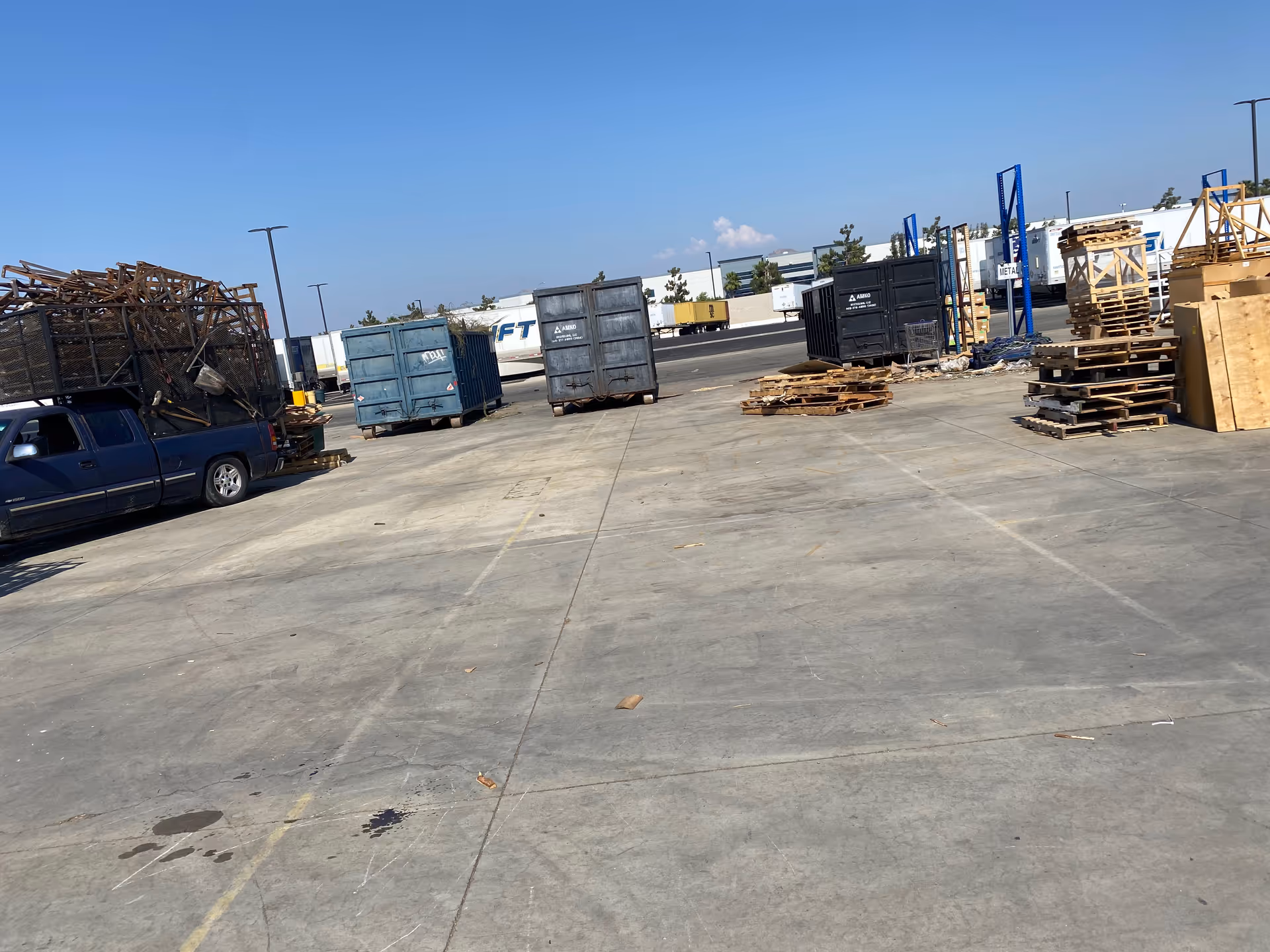 This wide outdoor industrial yard shows dumpsters, pallets, and parked vehicles.