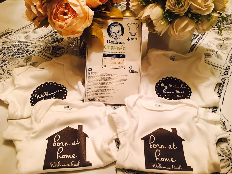 Three white baby onesies with a house motif, packaging, and flowers on a lace-draped table.