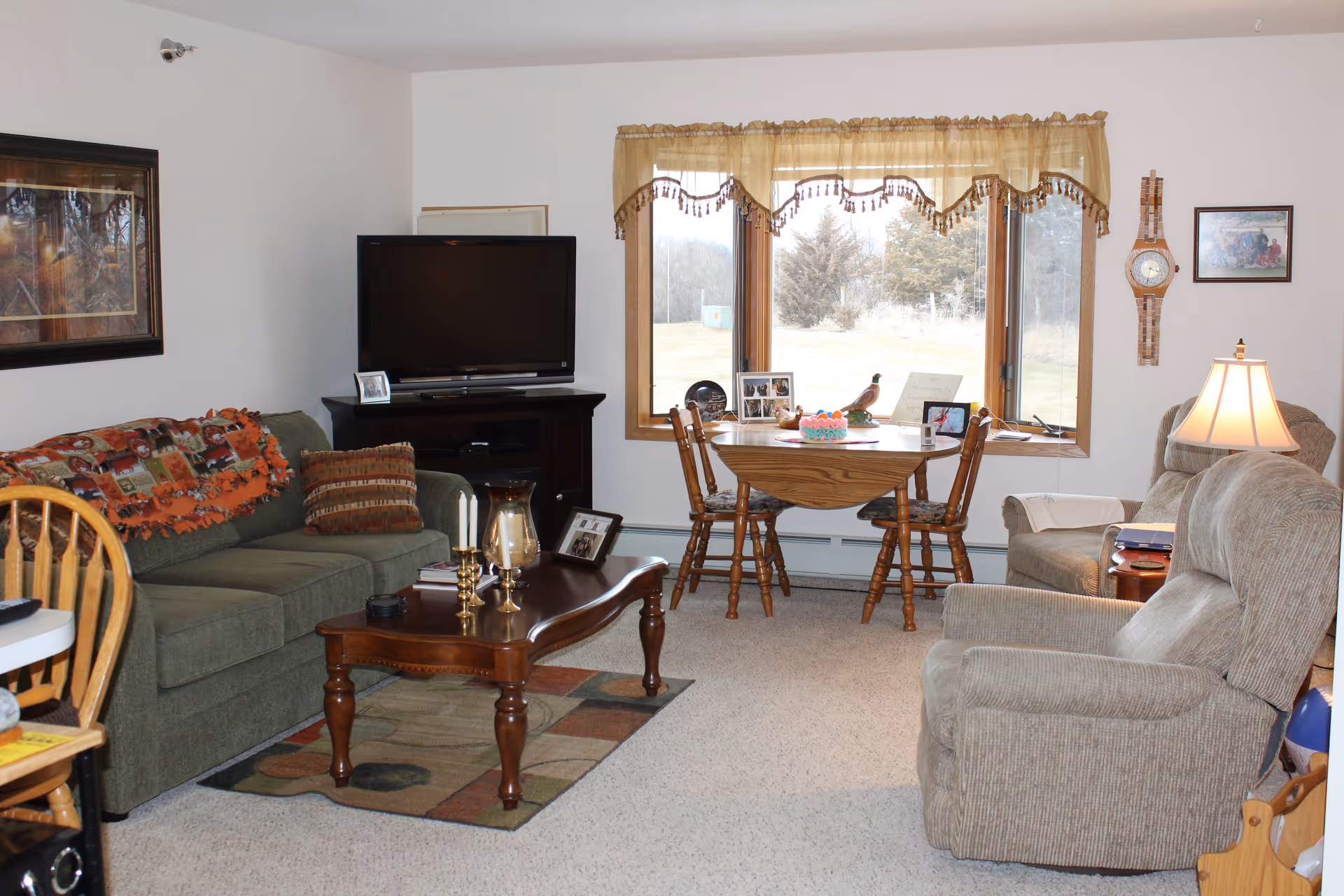 A cozy living room with a green sofa adorned with a colorful throw blanket and a striped pillow, a wooden coffee table with decorative items, a flat-screen TV on a dark wooden stand, two beige recliners, a wooden dining table with four chairs near a large window with sheer curtains, and a lamp on a side table. The room has light-colored carpet and walls with framed pictures and a wall clock.