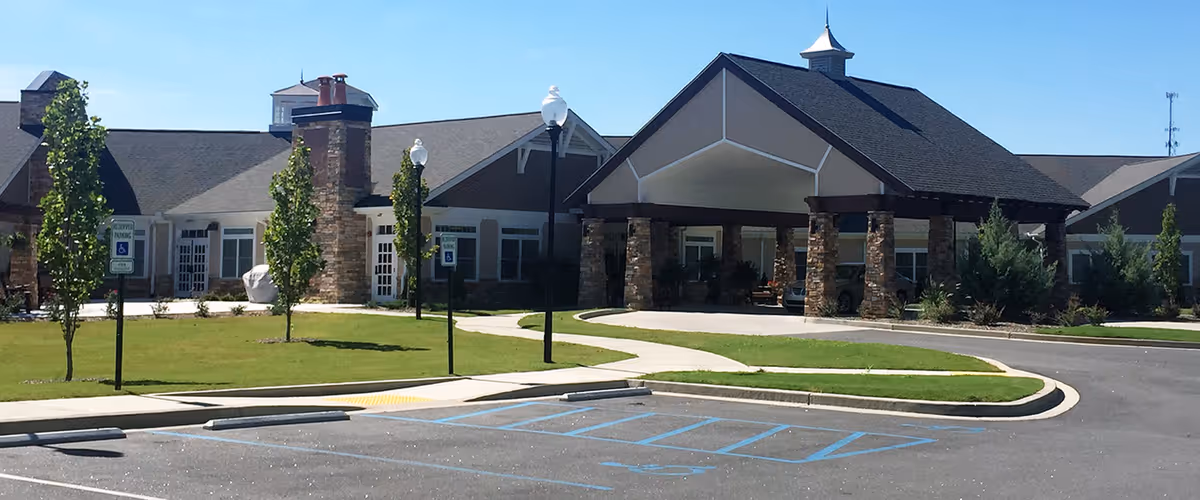 Exterior view of a single-story senior living facility building with a covered entrance supported by stone pillars, a driveway, parking spaces including handicapped spots, small trees, and a well-maintained lawn under a clear blue sky.