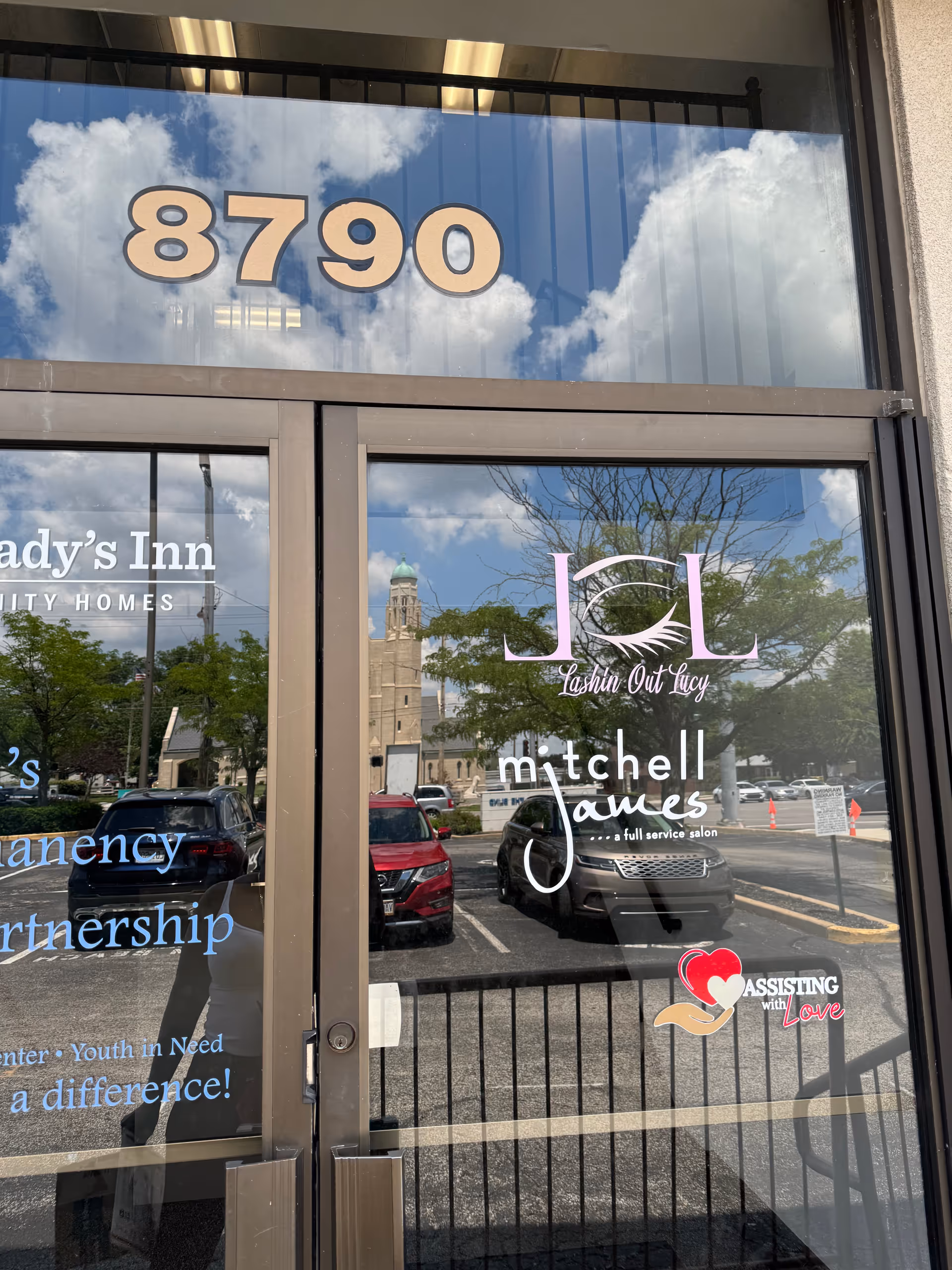 Glass storefront with 8790 and 'Assisting with Love' logo, reflecting parked cars outside.