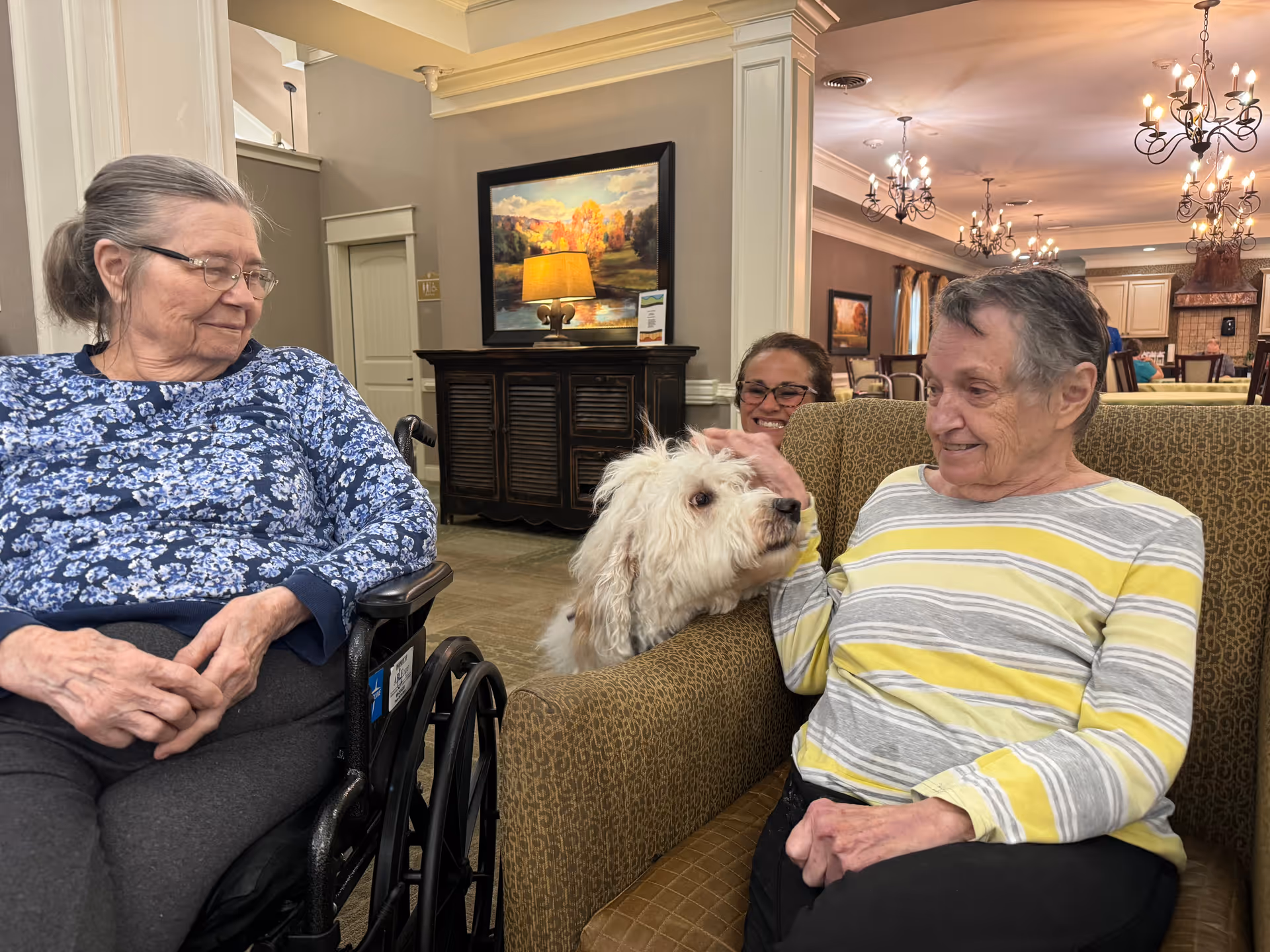 Two elderly women sitting indoors in a common area, one in a wheelchair wearing a blue floral top and the other on a patterned couch wearing a yellow and gray striped shirt, smiling at a white fluffy dog being petted by a third person partially visible behind the couch. The room has warm lighting, chandeliers, and framed artwork on the walls.