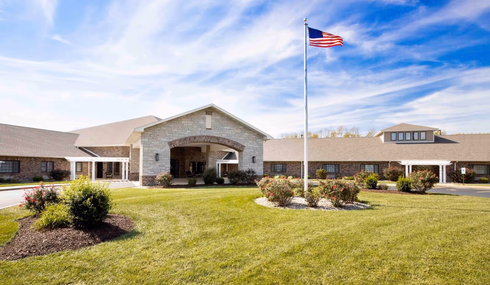 Exterior view of Cedarhurst Senior Living of Bethalto building with a well-maintained lawn, flower beds, and an American flag on a flagpole under a partly cloudy blue sky.
