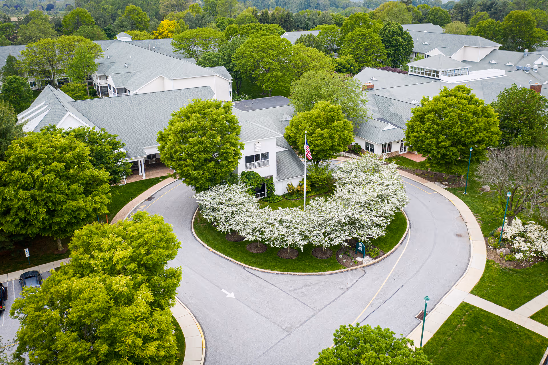 Aerial view of a senior living campus entrance with a circular driveway, flagpole, blooming shrubs and surrounding buildings.