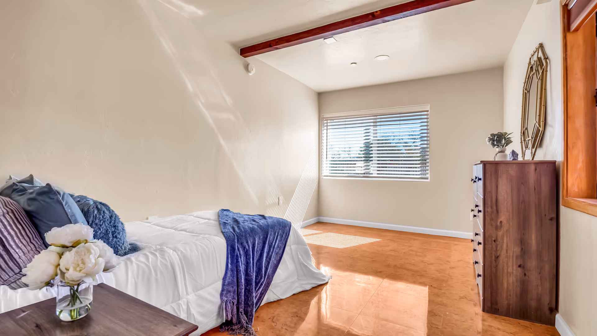 A tidy bedroom with a bed, dresser, plant decor, and a window with blinds.