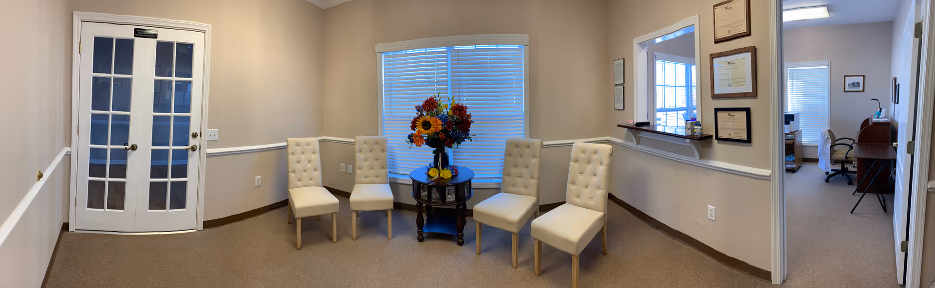 Beige-walled interior lounge with four white tufted chairs around a round table with flowers.