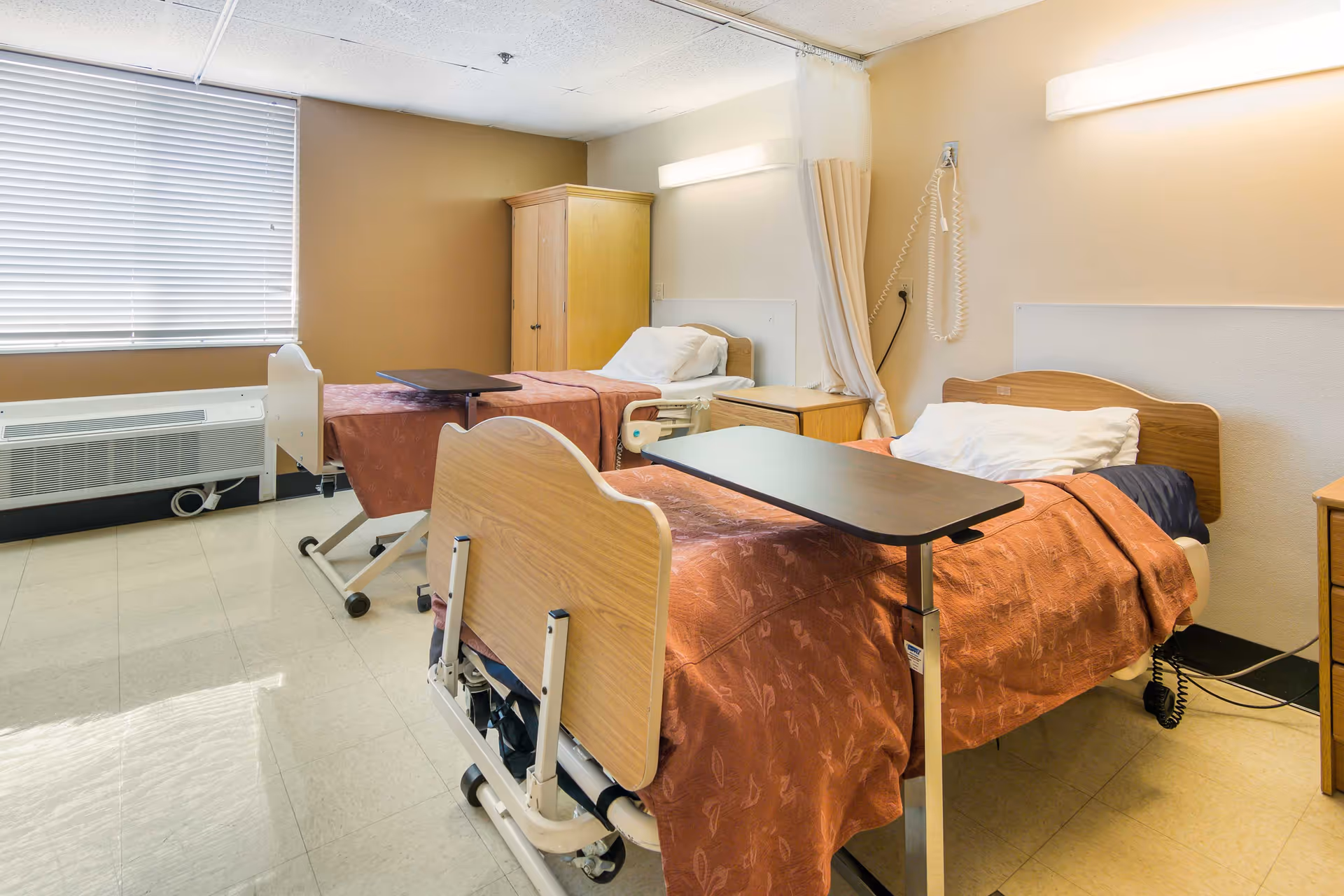 Two hospital beds with orange bedding in a shared patient room with desks and curtains.