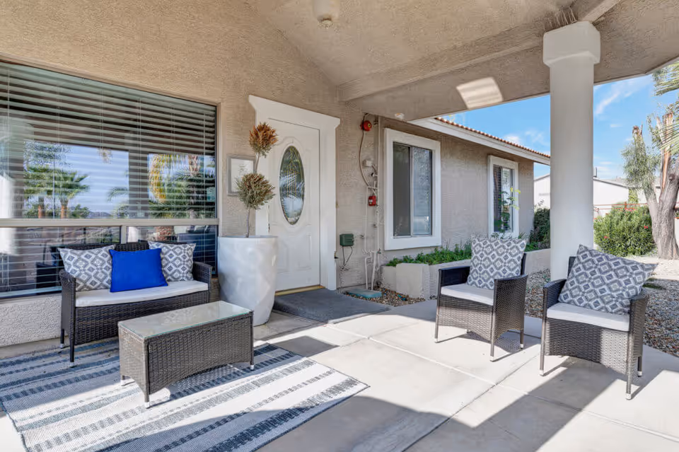 Outdoor covered patio area at Valencia at 19th 1 featuring a white front door with an oval glass window, a large window with blinds, a potted plant, a striped outdoor rug, a wicker loveseat with cushions and pillows, and two matching wicker chairs with patterned cushions. The patio is shaded by a roof supported by a column, and there are trees and shrubs visible in the background under a clear blue sky.