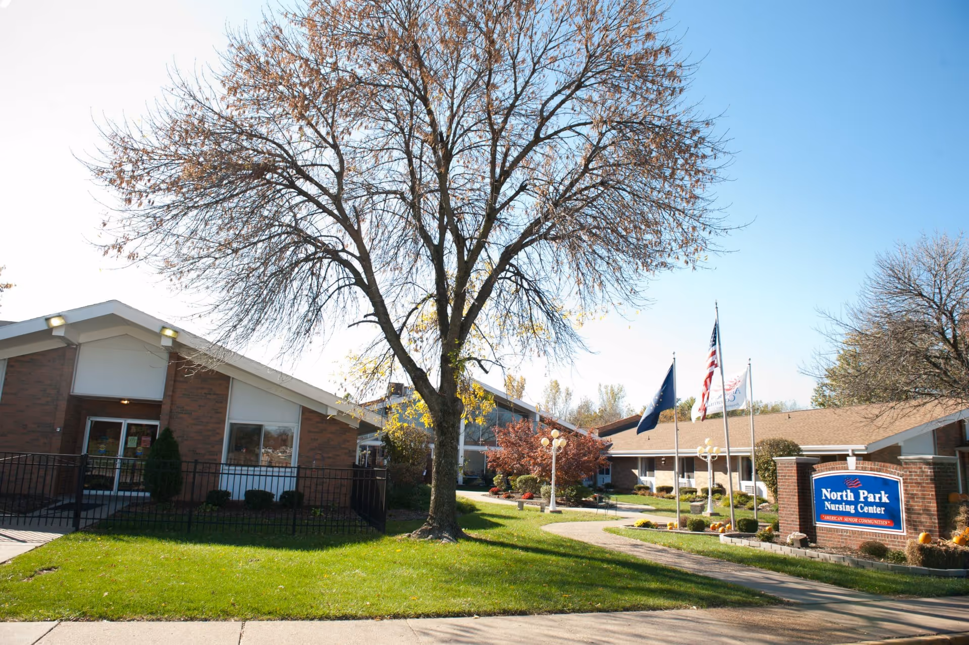 Front exterior of North Park Nursing Center with brick buildings, a large tree, flagpoles, walkway, and the facility sign.