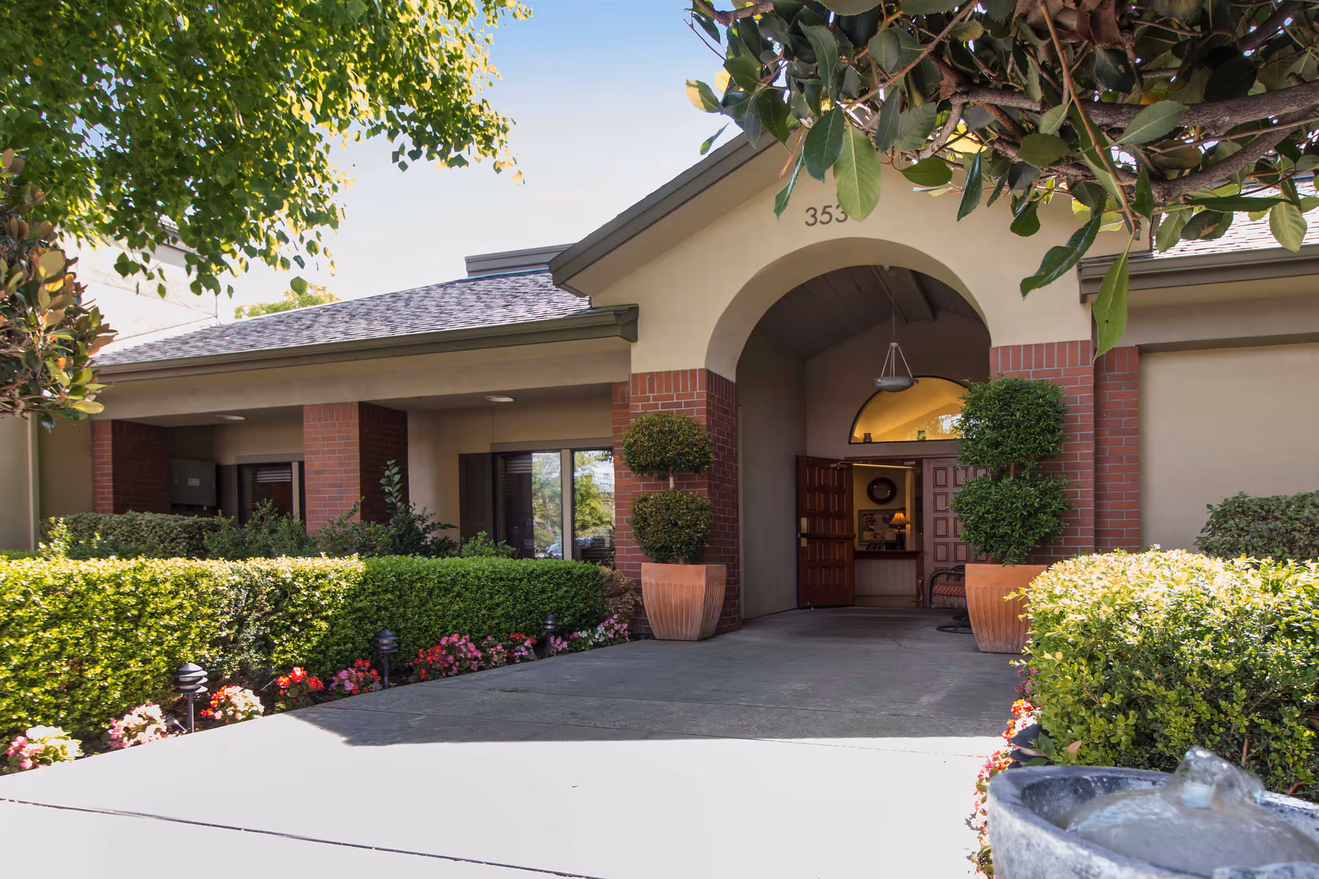 Front entrance of a single-story building with an arched doorway, potted topiaries, brick columns, and landscaped hedges and flowers.