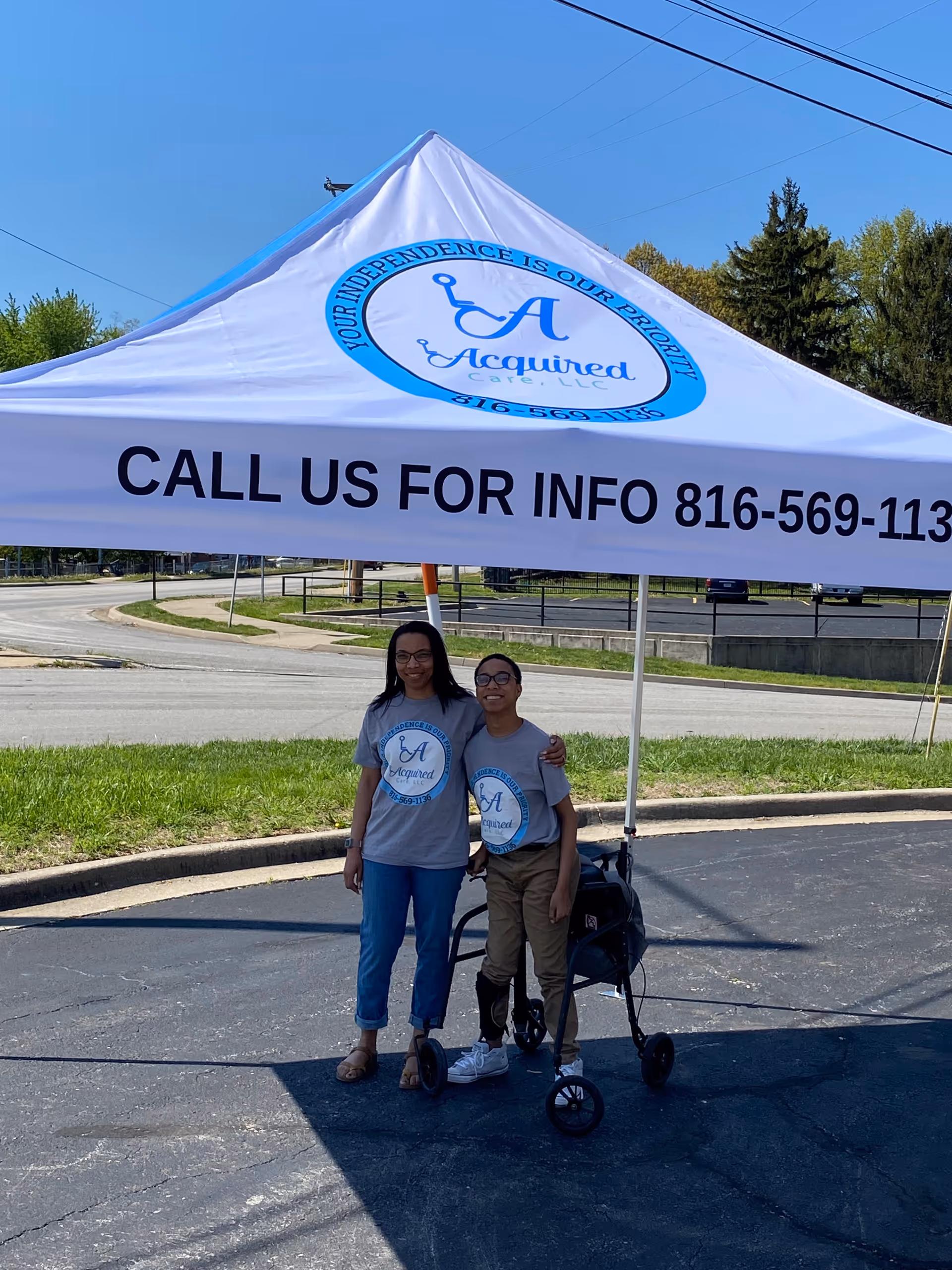 Two smiling individuals stand under an Acquired Care tent in a parking lot.