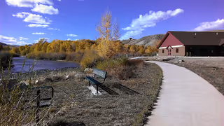 A paved walkway curves past benches along a riverside with autumn trees and a building under a blue sky.