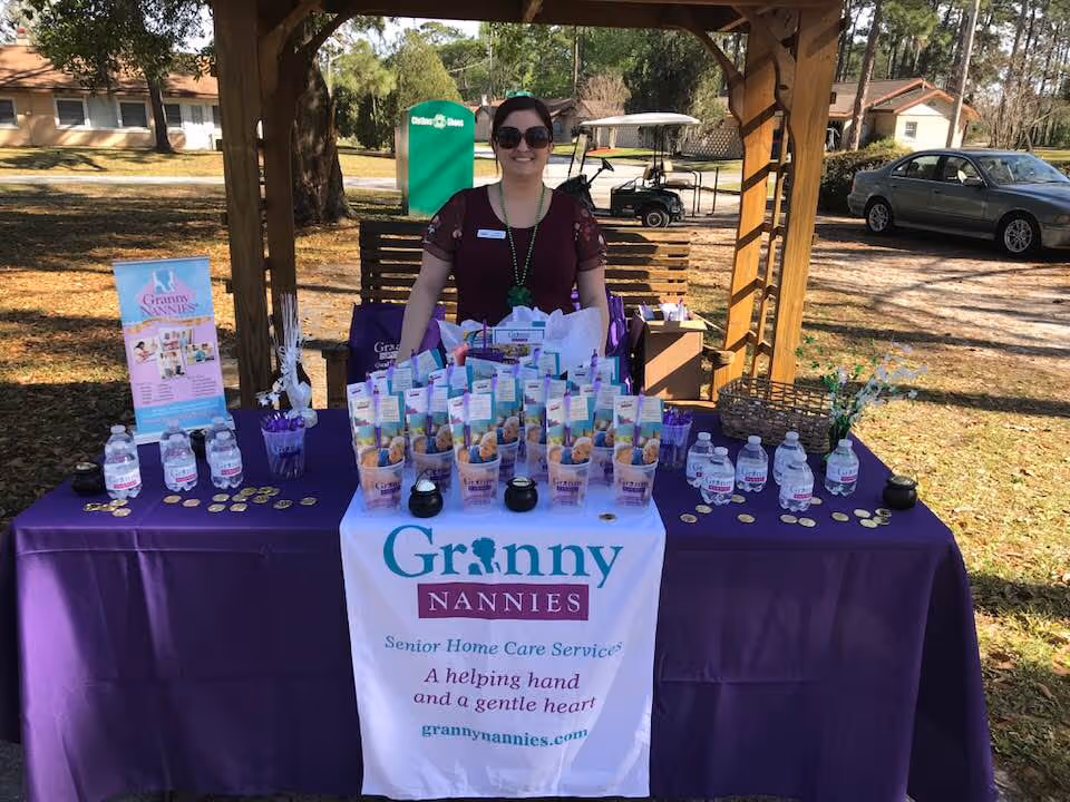 Staff member stands behind a purple Granny Nannies table at an outdoor booth.