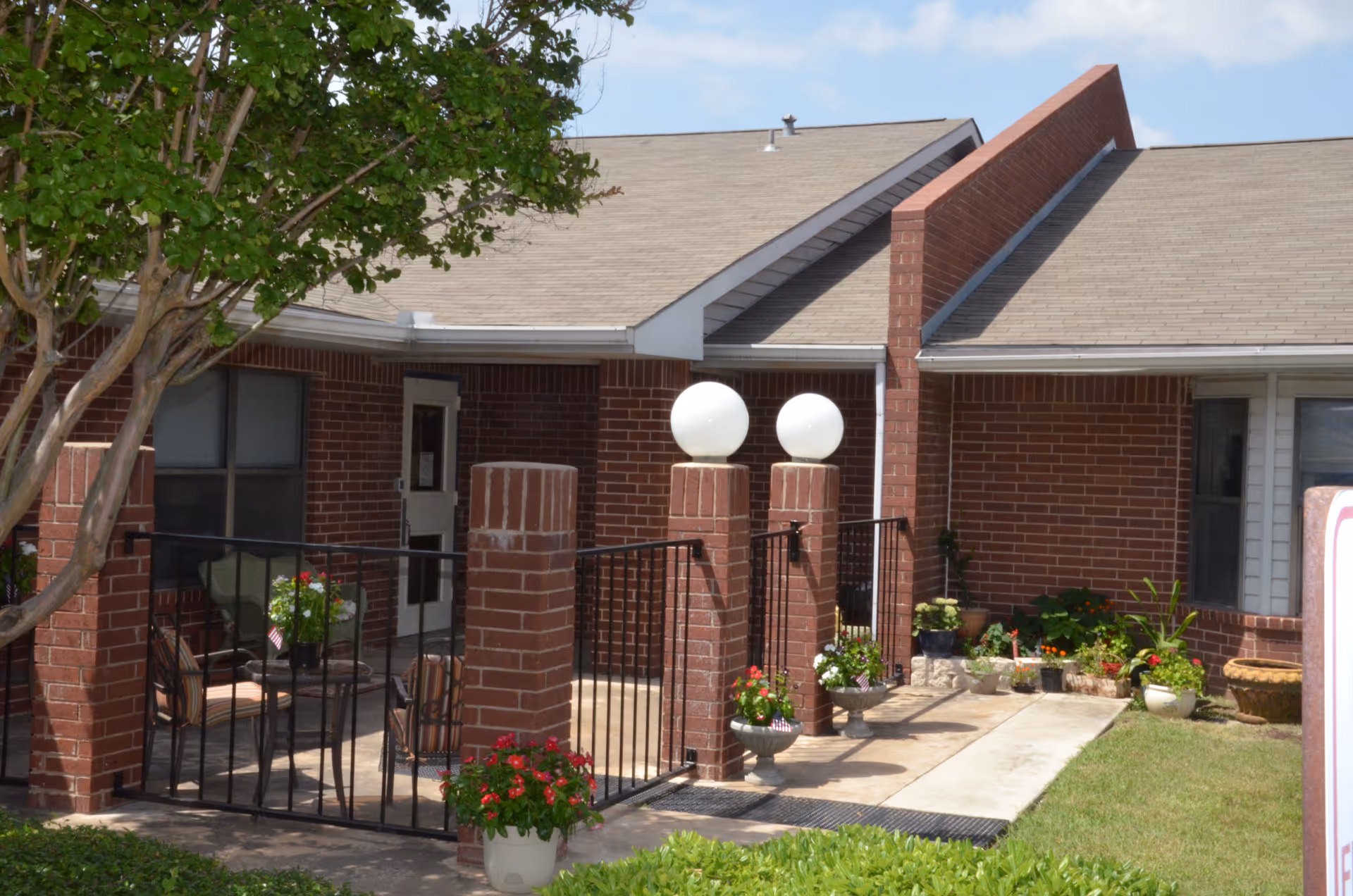 Outdoor patio area of a brick building with a small fenced seating area containing chairs and a table with potted plants. There are two brick pillars topped with round white lights, additional potted plants along the walkway, and a tree partially visible on the left side.
