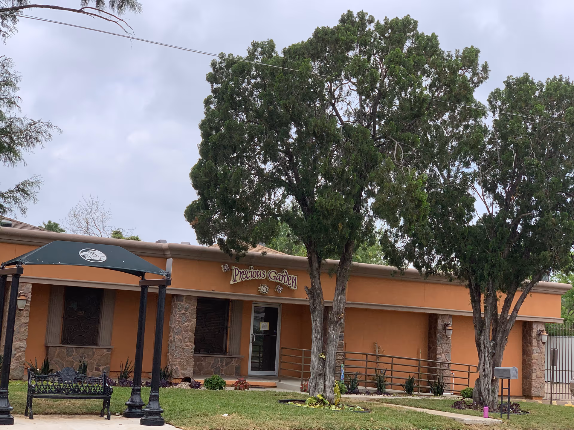 Orange single-story building with Precious Garden sign, trees, bench, and entrance.