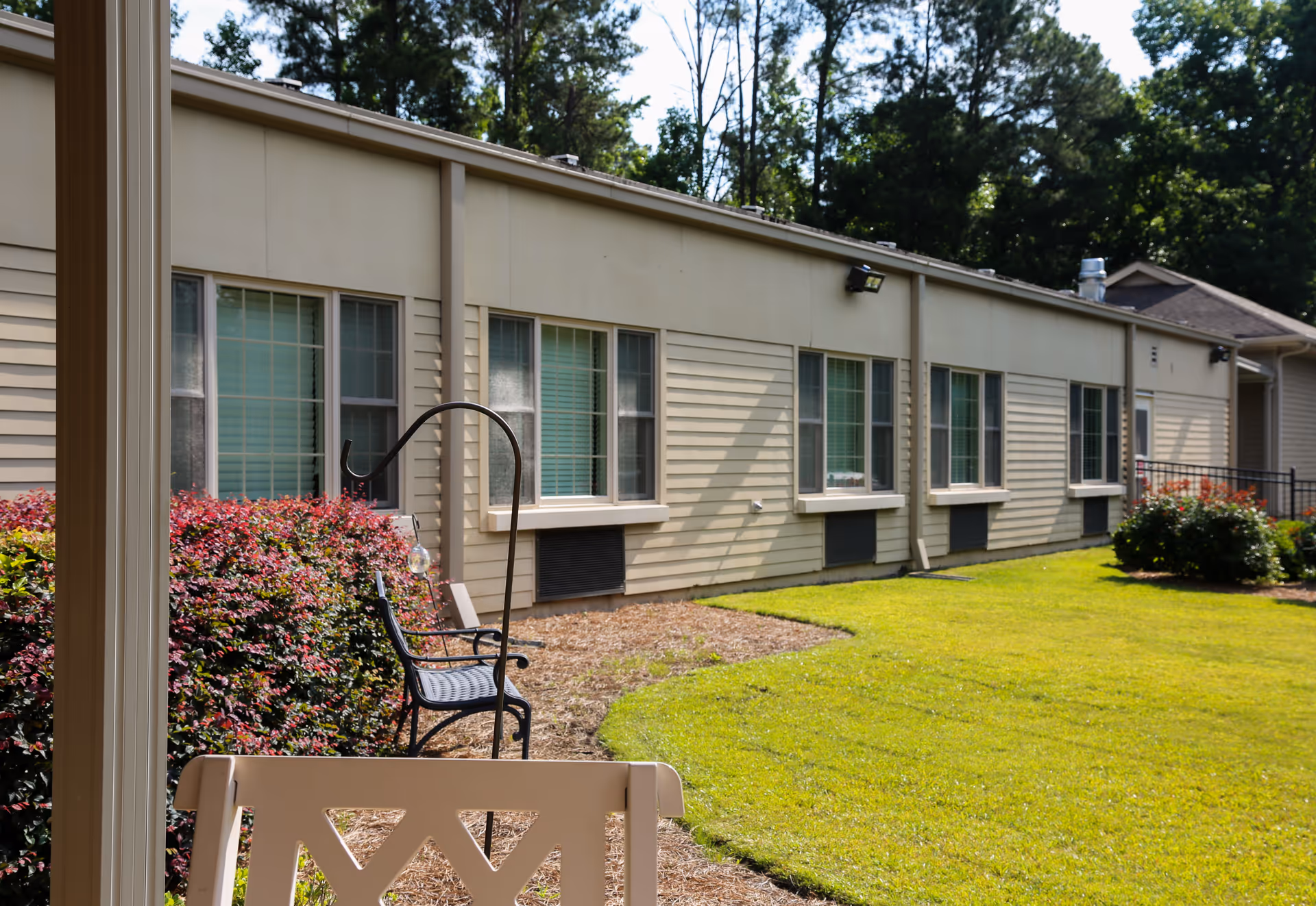 View of the exterior side of a single-story building with multiple windows, surrounded by green grass, bushes, and trees. There is a black metal chair and a beige bench in the foreground, with a garden area and a clear sky above.